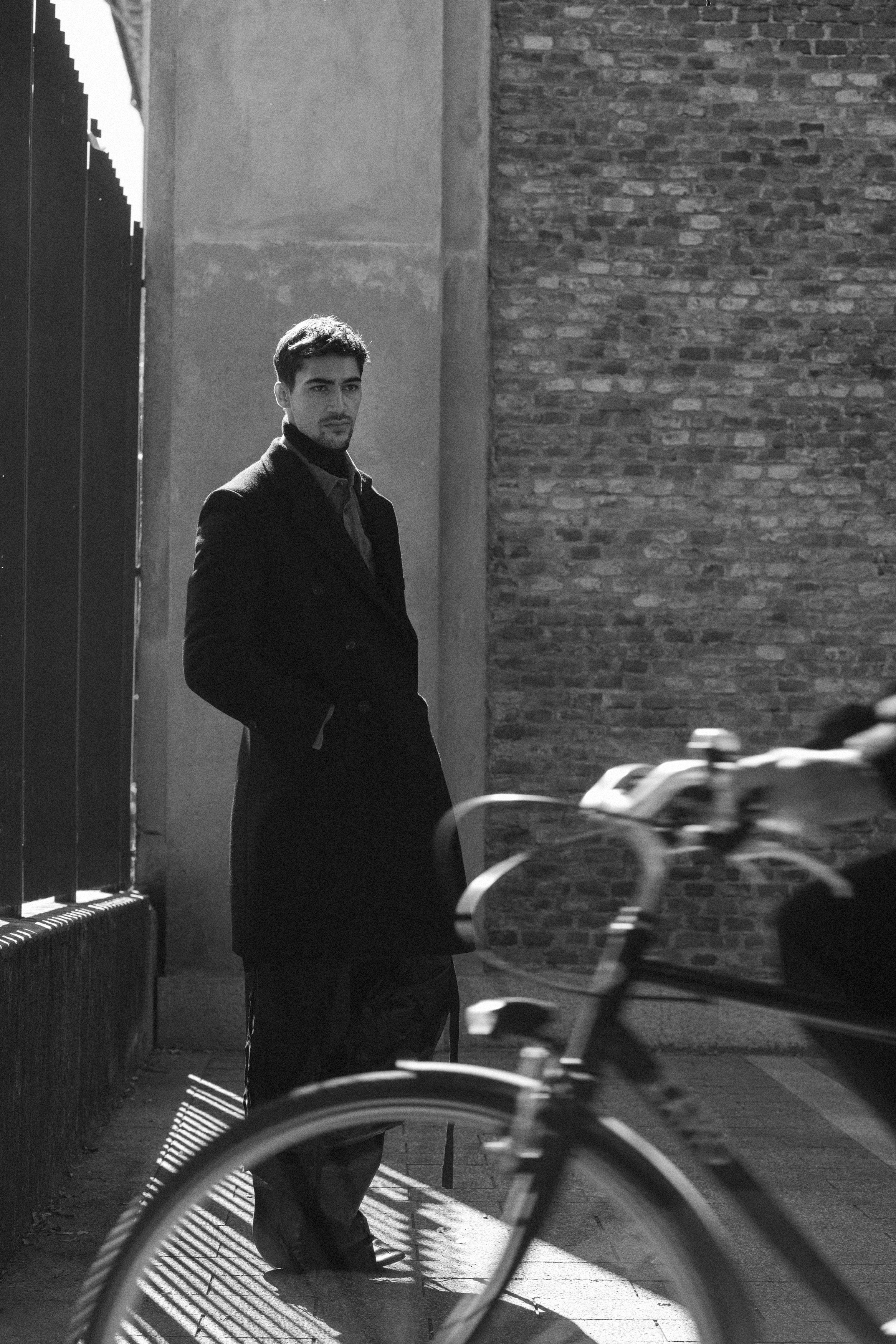 A young man in a dark coat and turtleneck standing on a sidewalk at night, next to a brick wall and a metal fence, with a bicycle partially visible in the foreground.