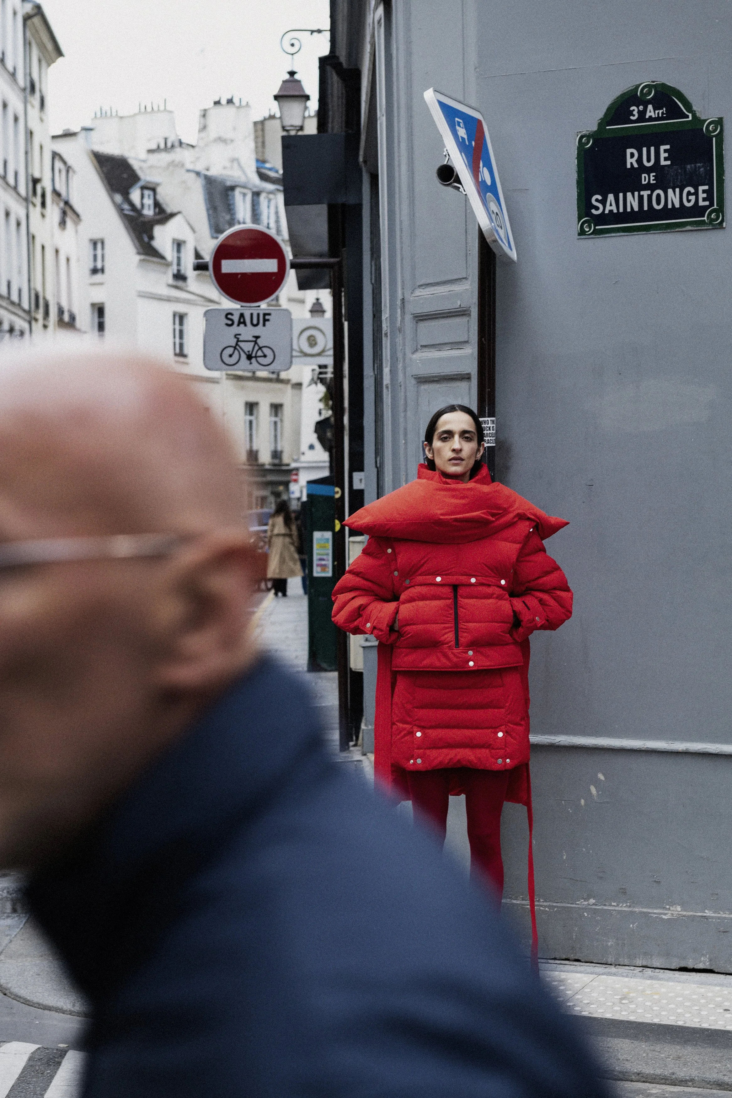 A woman in a red puffy jacket with a large collar stands on a street corner in Paris, France, next to a gray wall with street signs, one reading 'Rue de Saintonge.' A blurred person in the foreground walks past, while other pedestrians walk along the