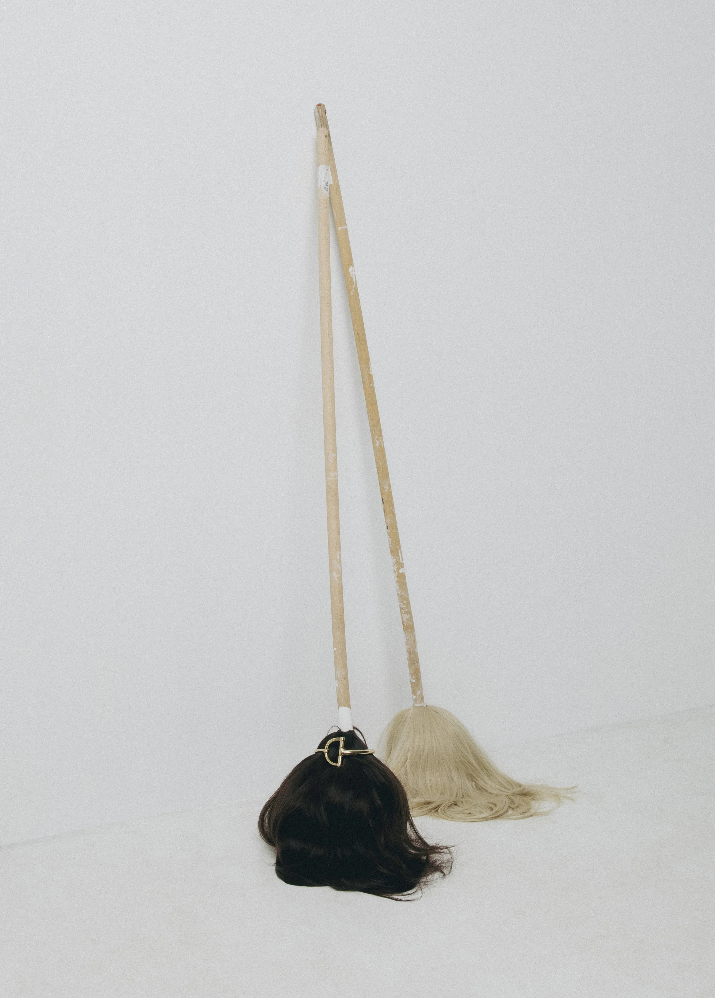 Two wigs, one black and one blonde, are resting on the floor against a white wall, with a broom leaning on the wall behind them.