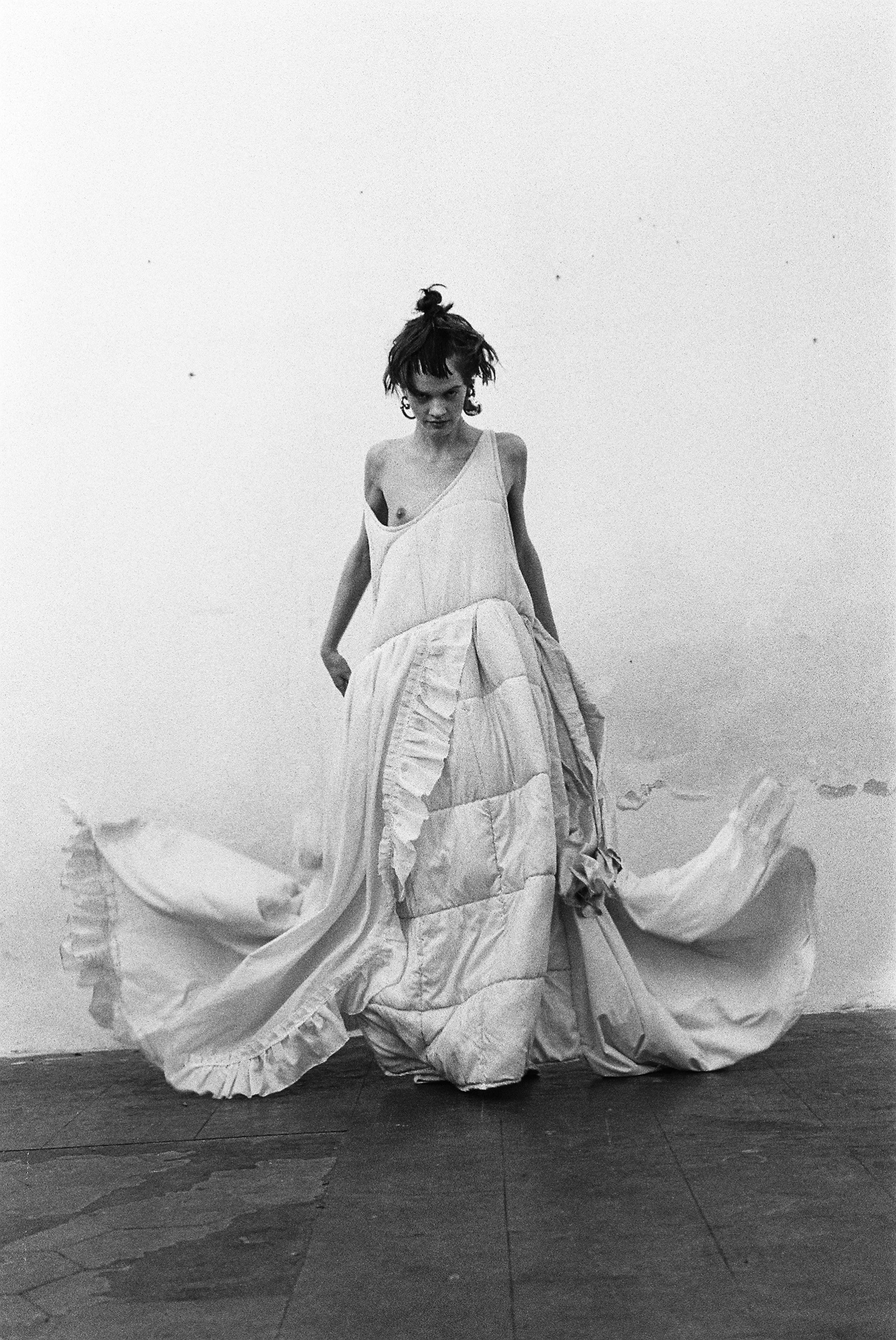 A woman in a long, flowing dress standing against a plain wall, looking down with a serious expression.