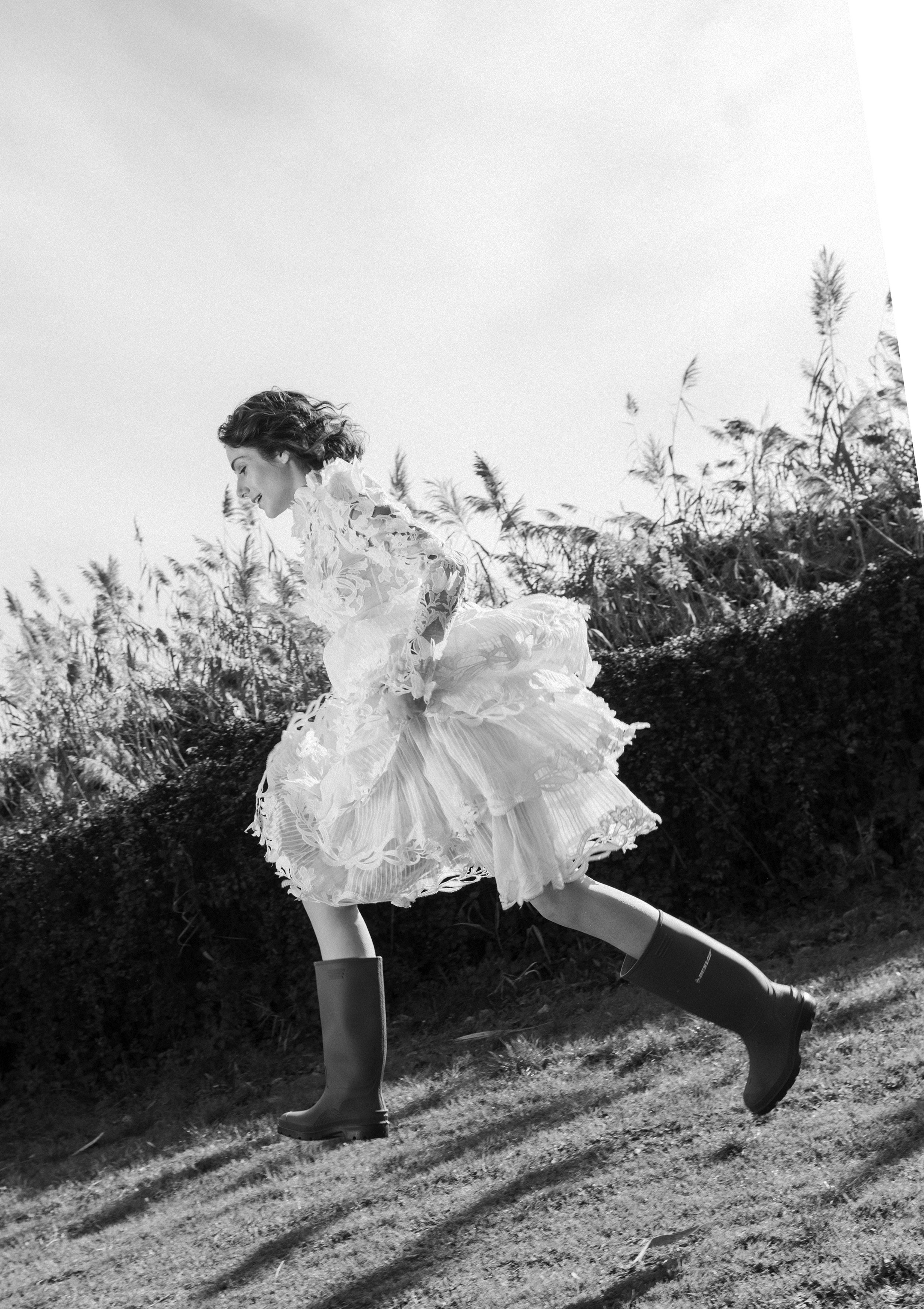 A woman in a floral dress and rubber boots running outdoors on grass with tall plants in the background, captured in black and white.