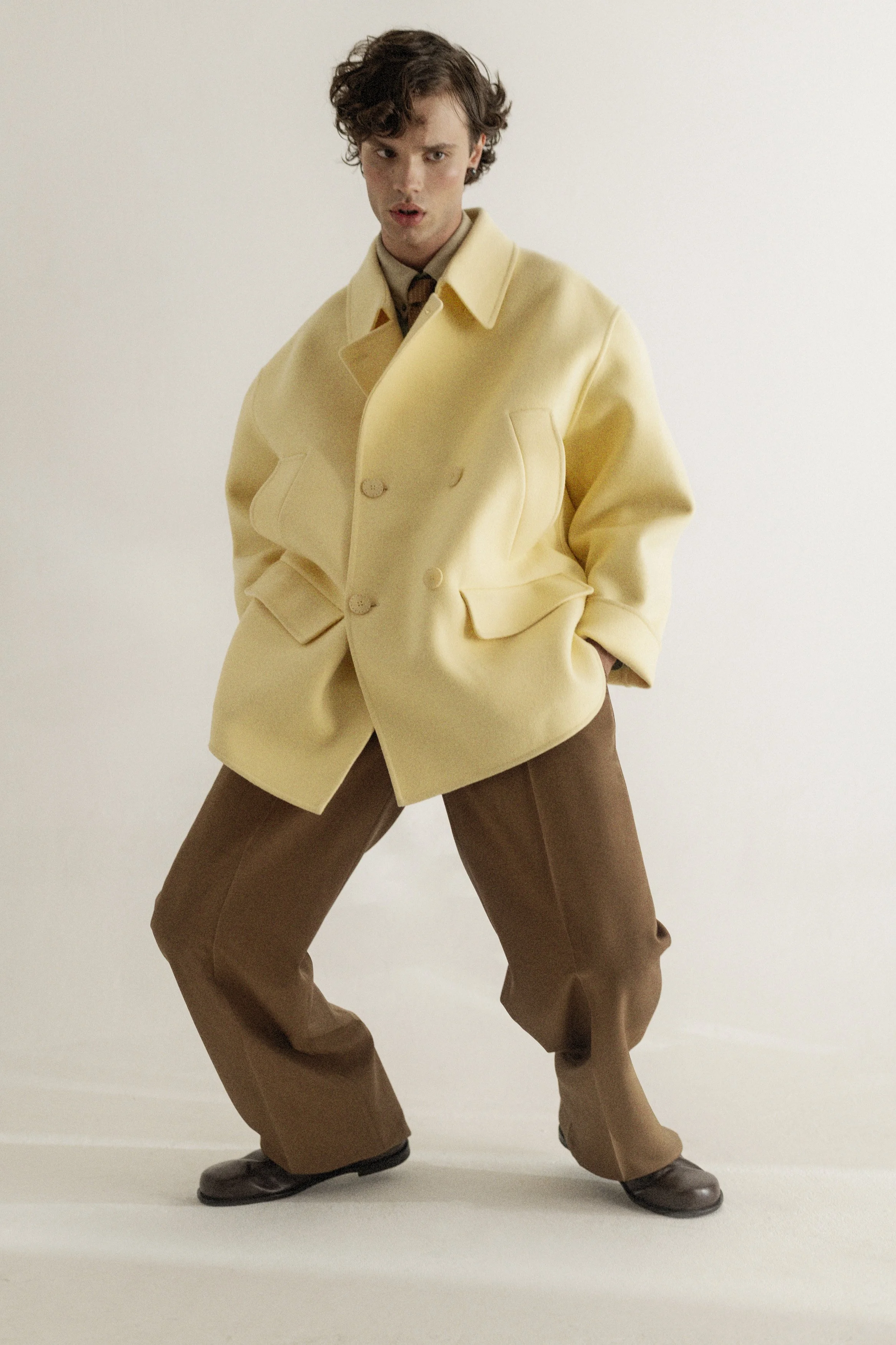 A young man with curly hair wearing a large beige coat, brown pants, and dark shoes, standing with a wide stance against a plain background.
