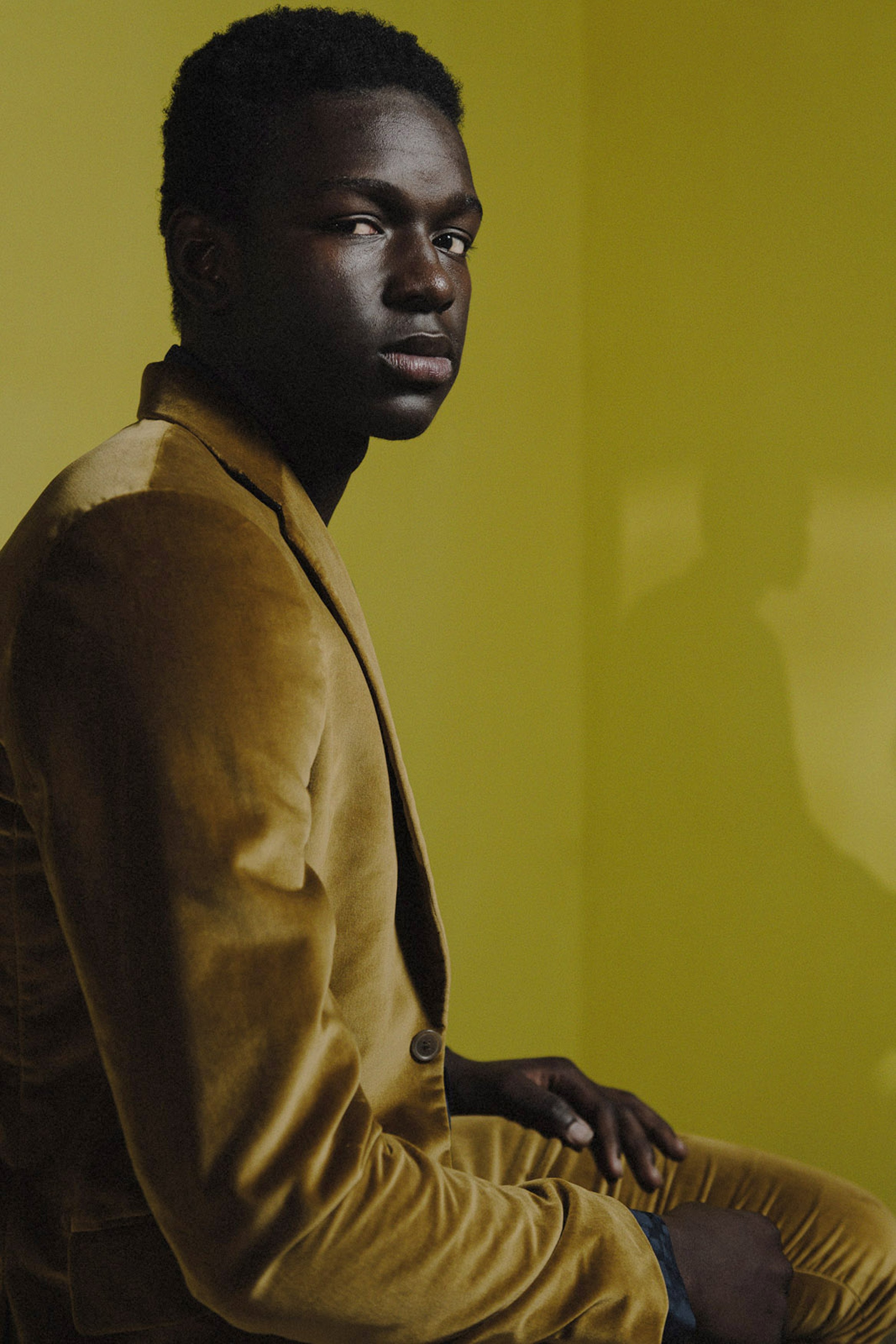 A dark-skinned man in a brown velvet suit sits against a yellow background, looking directly at the camera with a serious expression.