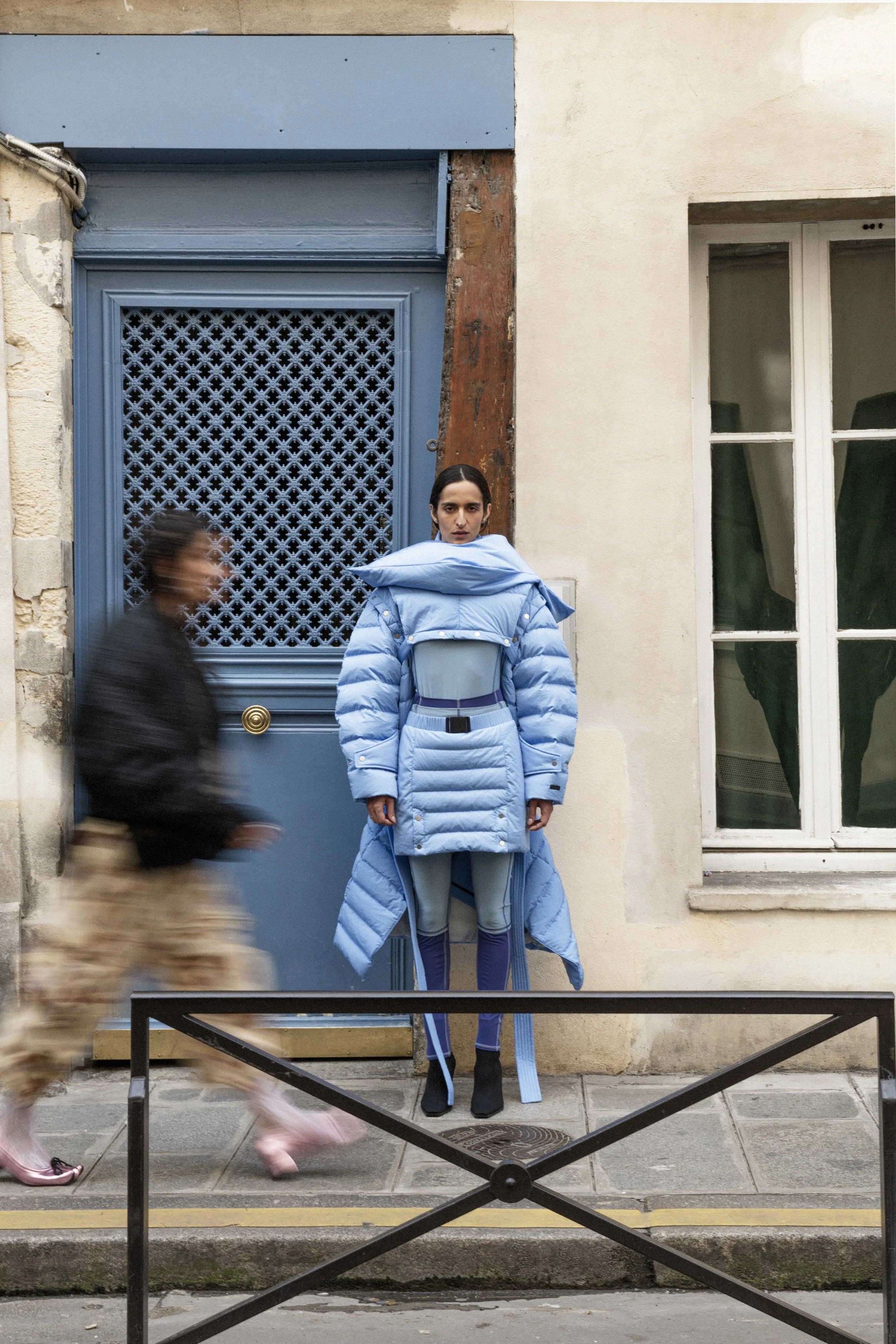 A woman standing against a beige wall and blue door, dressed in an elaborate blue puffer coat and matching gear, with a serious expression, while a blurred person walks past in front.