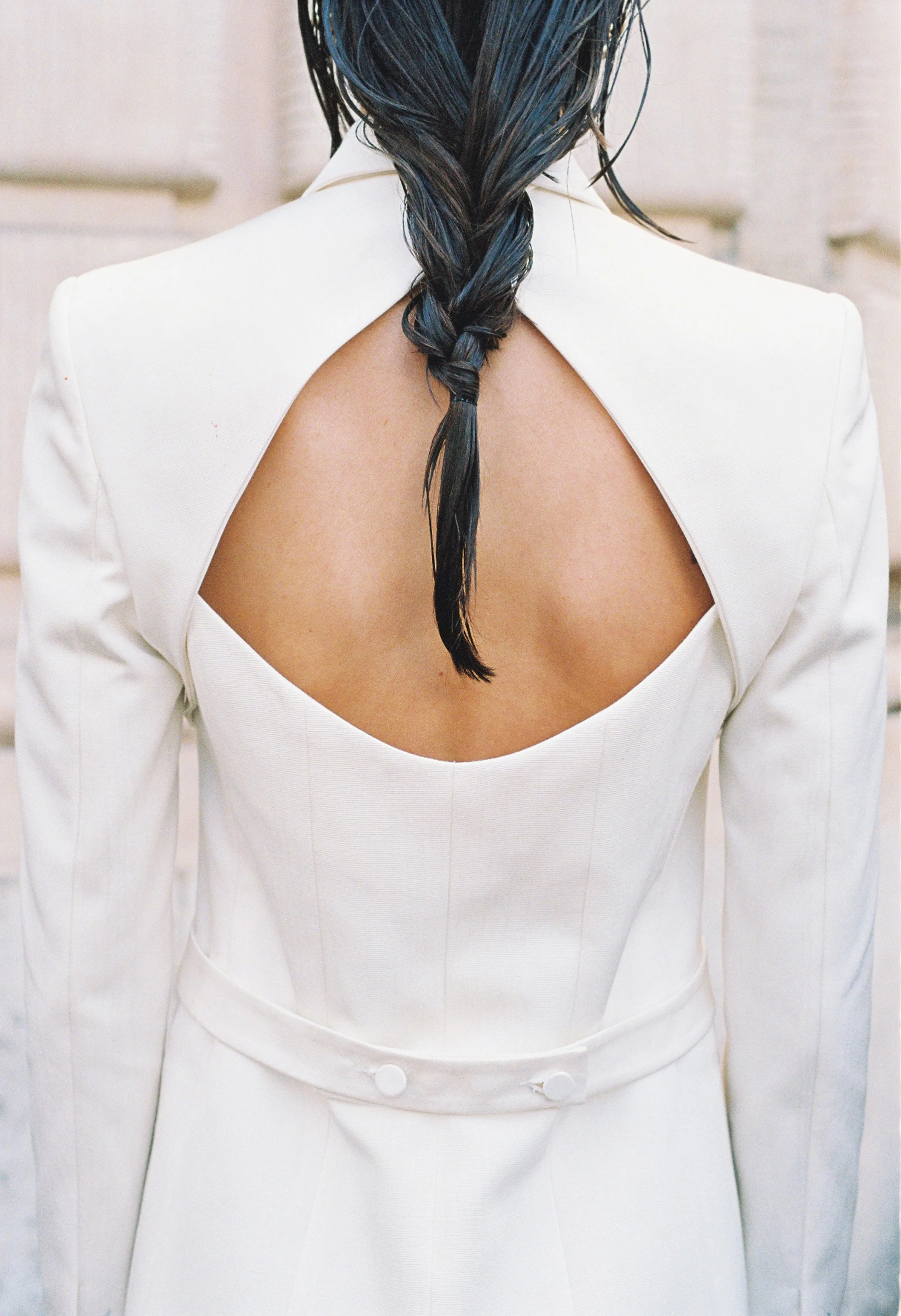 A woman wearing a white blazer with an open back design, revealing her upper back and shoulders, and her dark, wet hair styled in a braid.