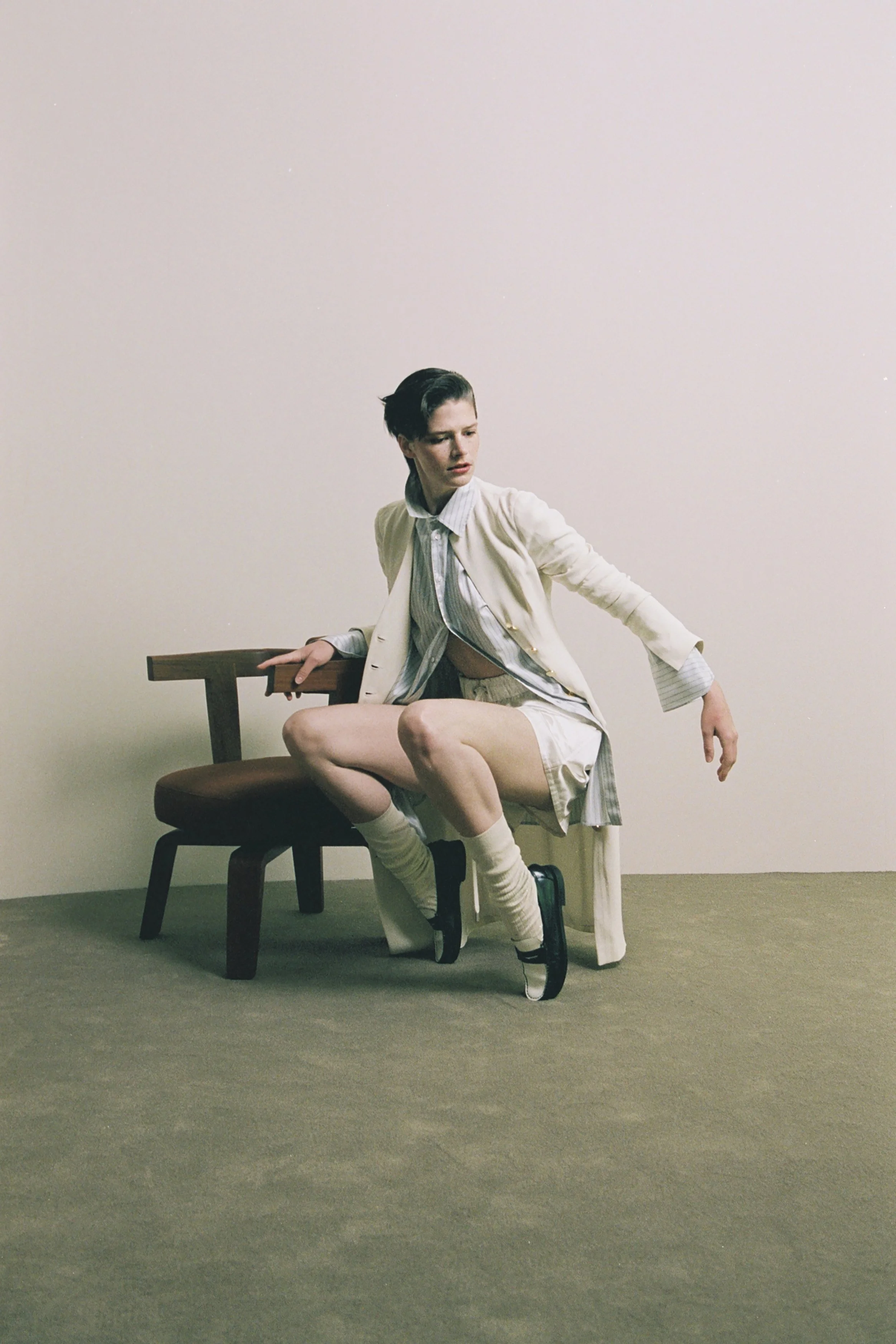 A woman with short dark hair sitting on a wooden chair, wearing a beige coat, a white shirt, and shorts, with one leg crossed over the other, in a minimalist room with a plain white wall and green carpet.