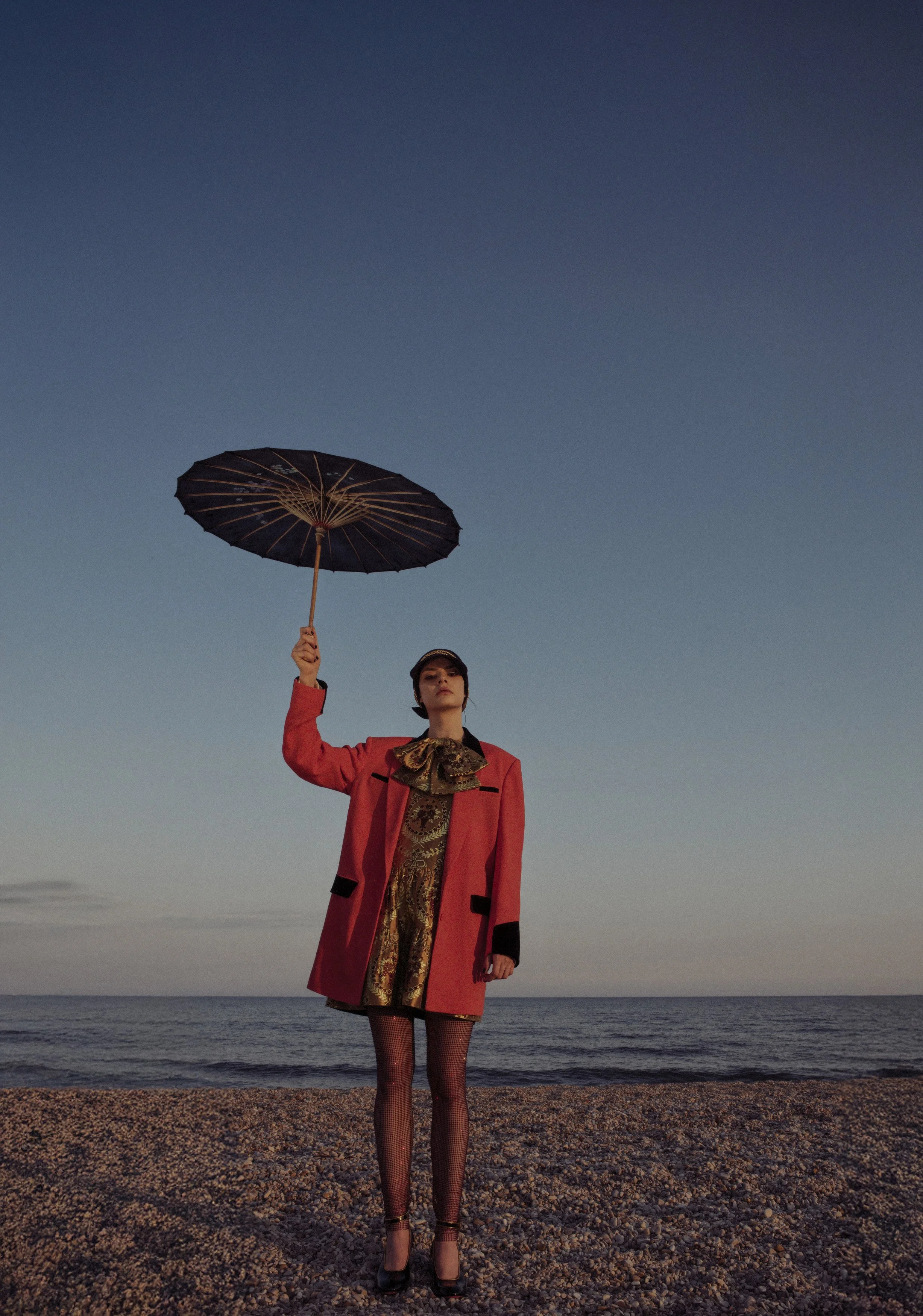 A woman in a vibrant red coat and ornate gold dress stands on a pebble beach holding a black umbrella, with the ocean and a clear sky in the background during sunset or twilight.