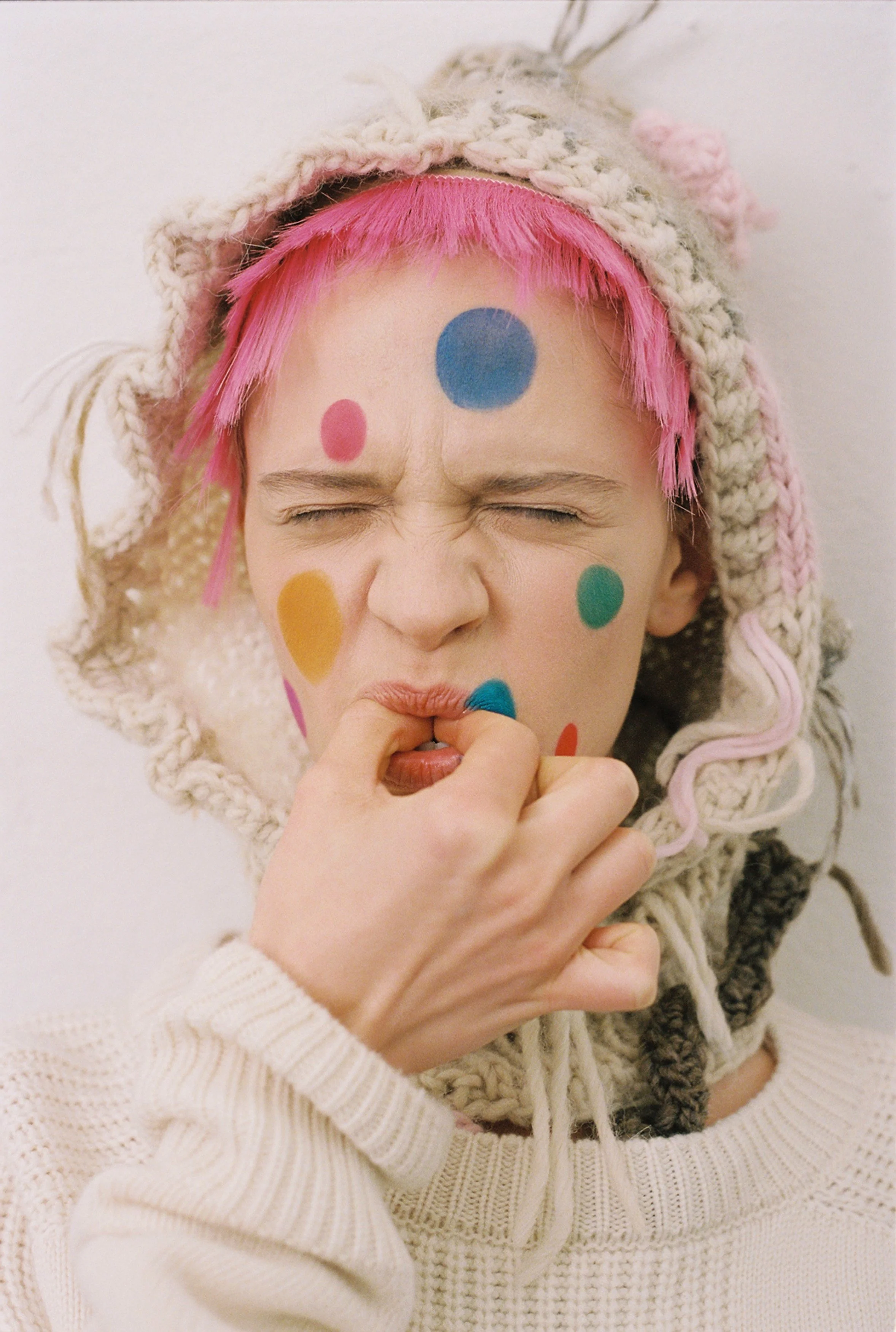 Person with pink hair, wearing a knitted hat, with colorful dots painted on face, biting finger, expressing distress or discomfort.