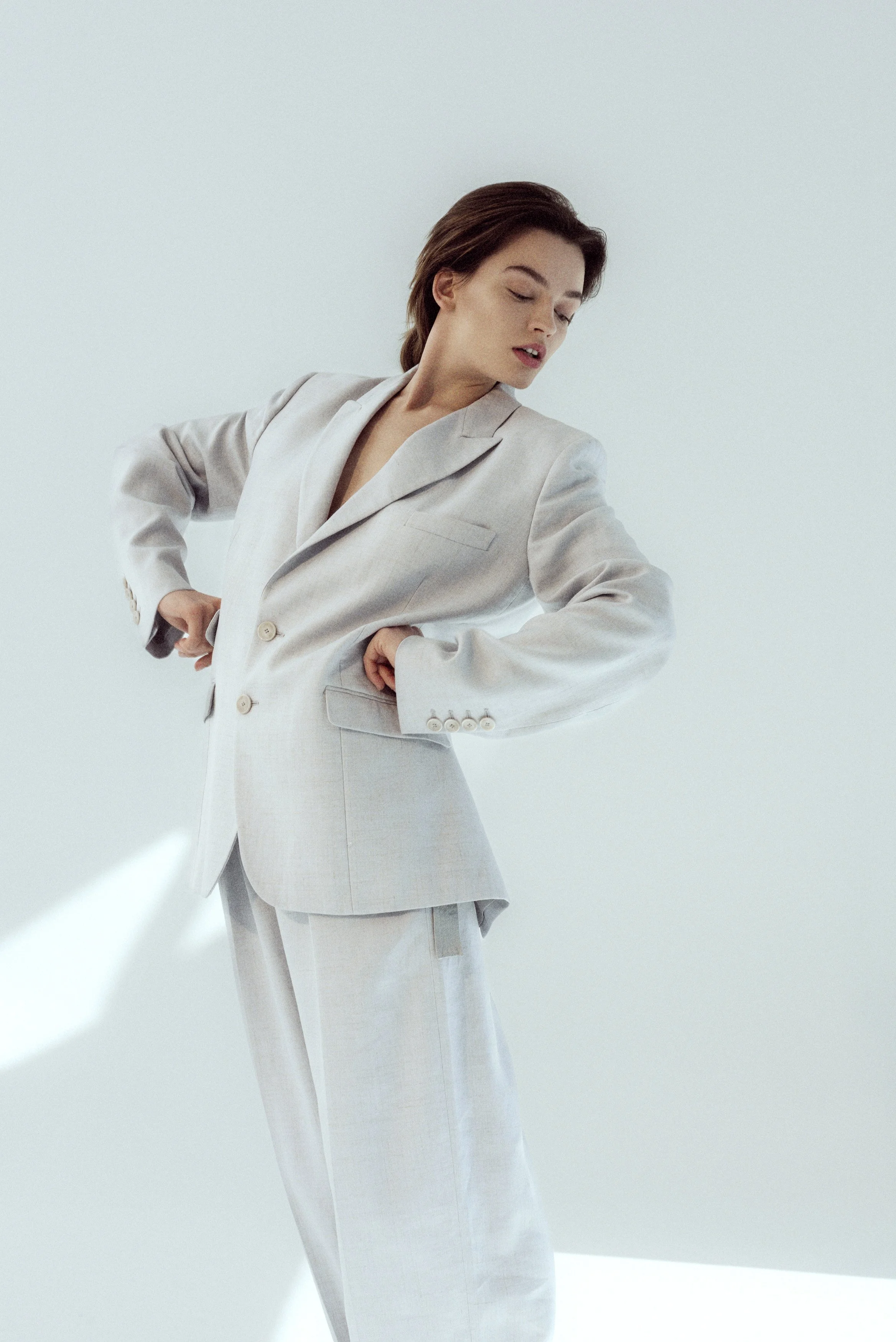 Woman wearing a light gray suit, standing against a plain light background, looking down with her hands on her hips.
