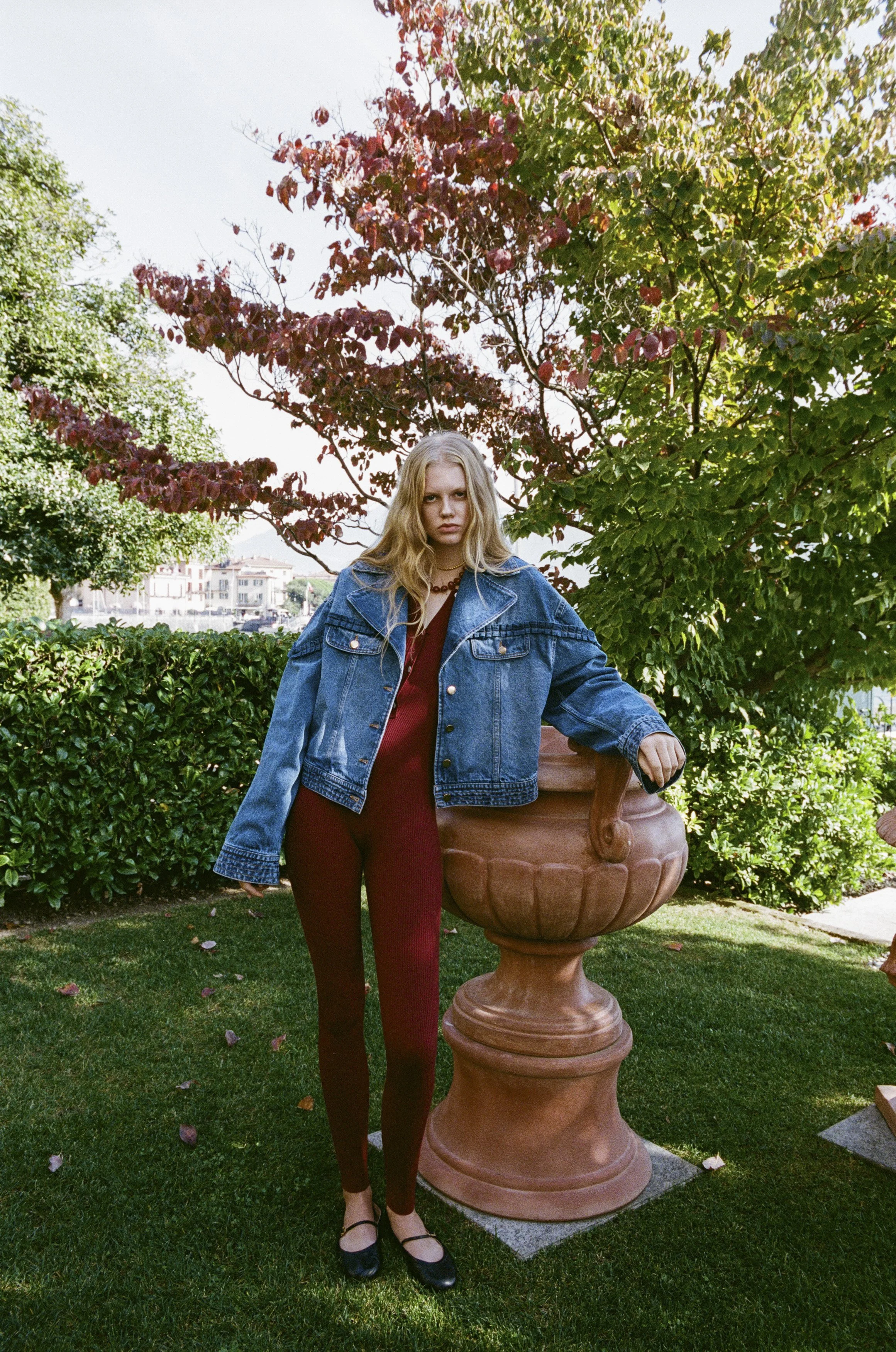 A young woman with blonde hair standing outdoors near a large decorative planter, surrounded by green bushes and trees with red and green leaves, wearing a denim jacket, red outfit, and black shoes.