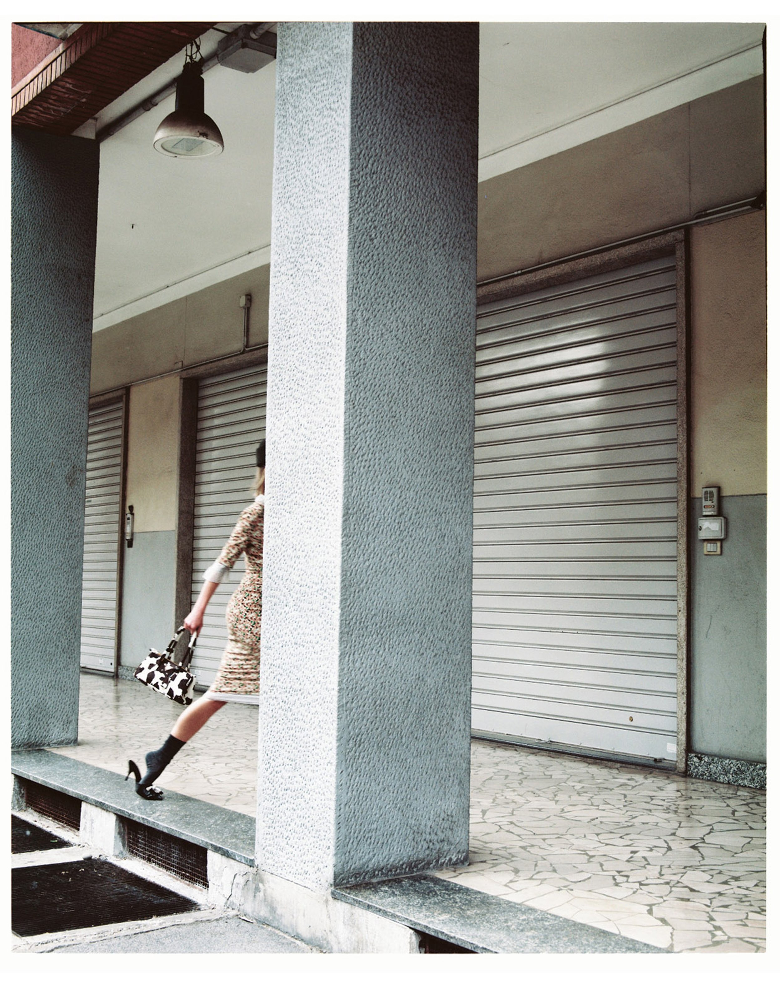 A woman in a patterned dress, high heels, and black socks is walking past closed store shutters in an urban setting, carrying a cowprint handbag.