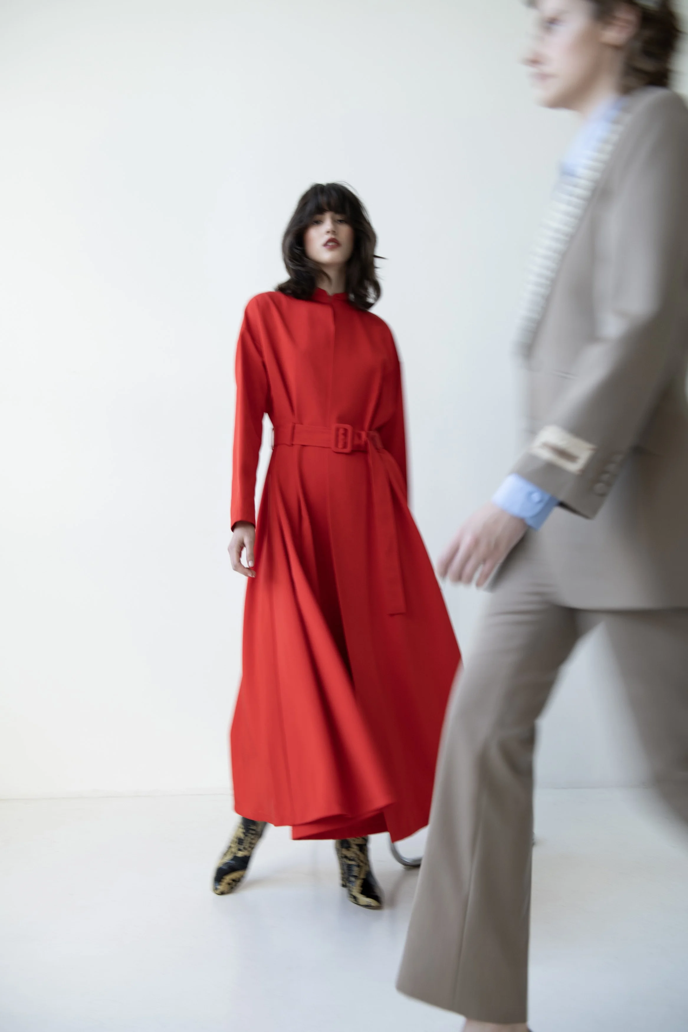 A woman wearing a bright red dress standing against a plain white background, with another person in a beige suit partially visible in the foreground.
