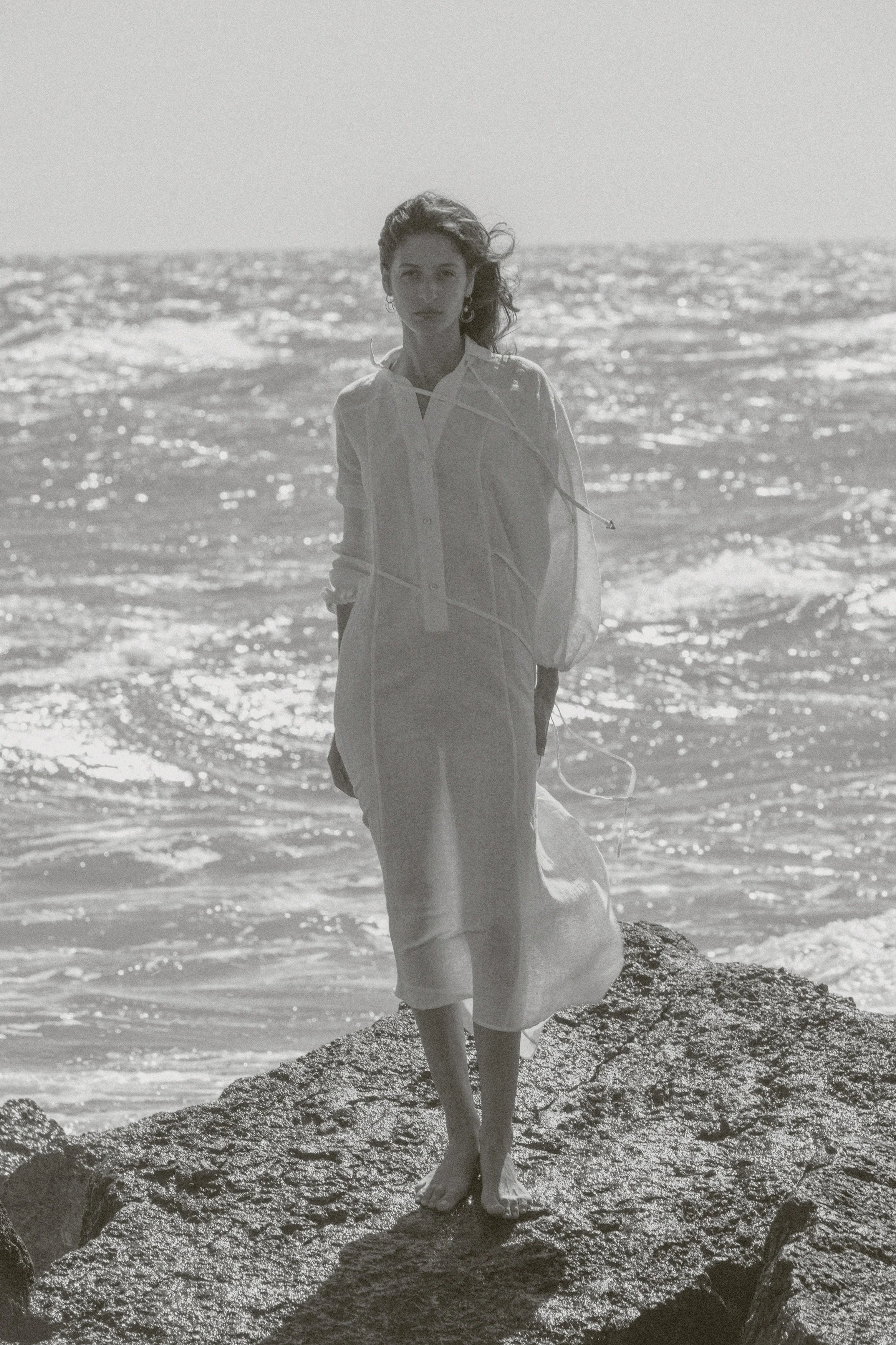 A woman standing barefoot on a rocky shoreline, facing the camera, with the ocean in the background and sunlight reflecting on the water.