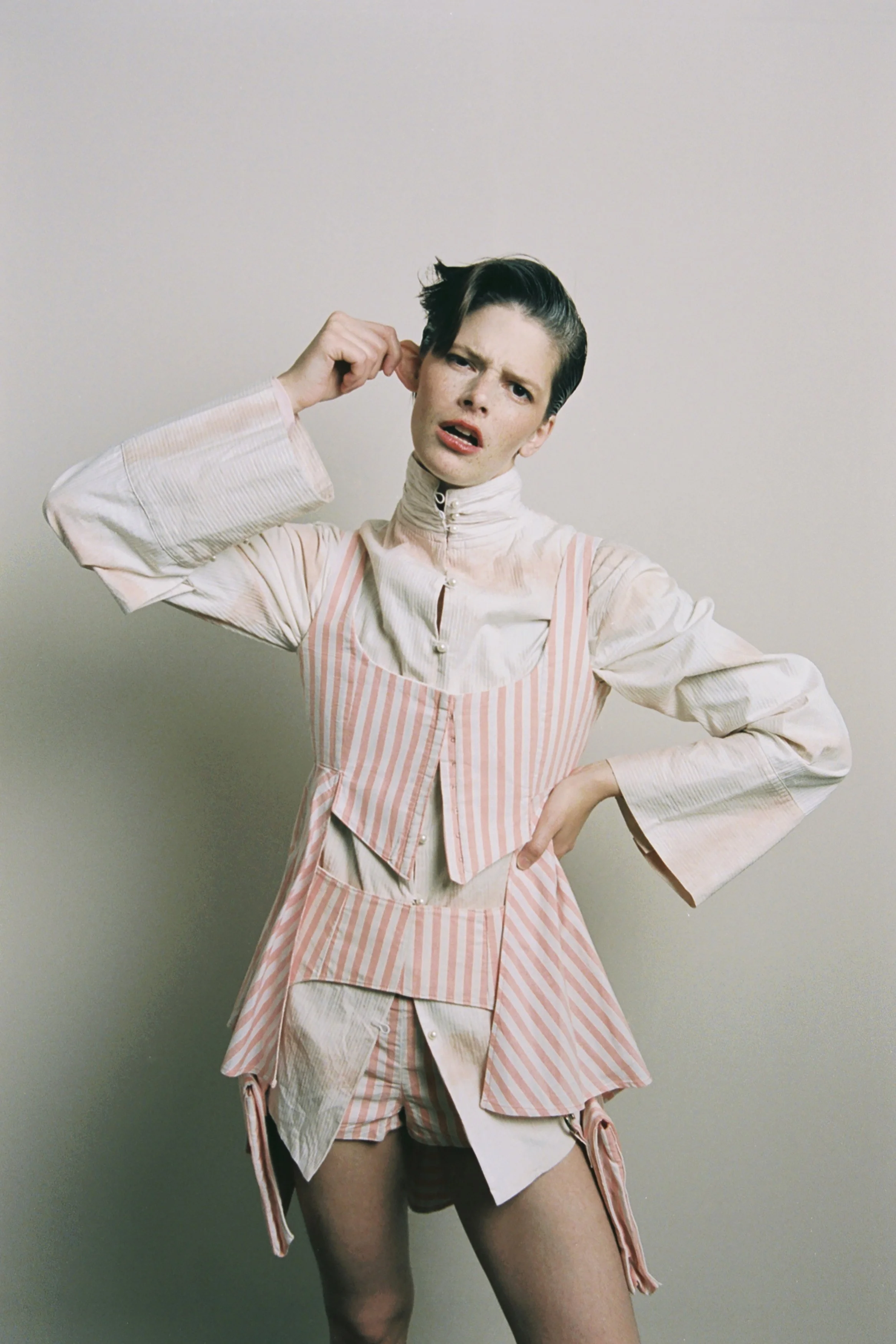Model with short dark hair wearing a beige and pink striped layered outfit, scratching their head with a confused or annoyed expression.