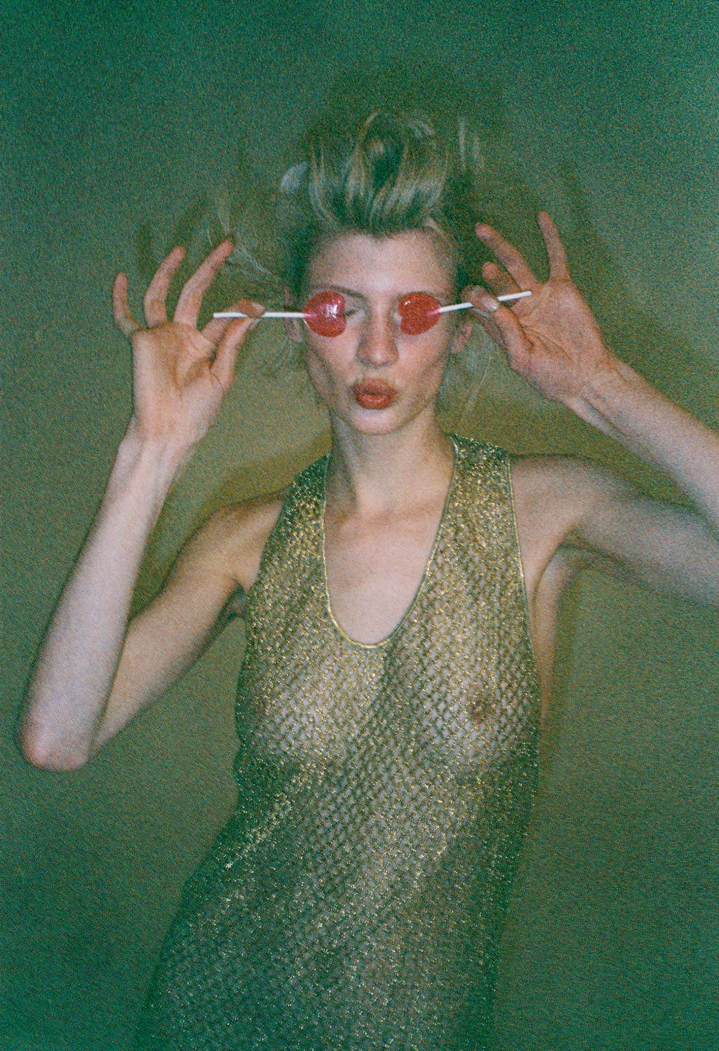 A woman with short gray hair lying on the ground, holding two heart-shaped lollipops over her eyes, wearing a sparkly gold sleeveless dress.