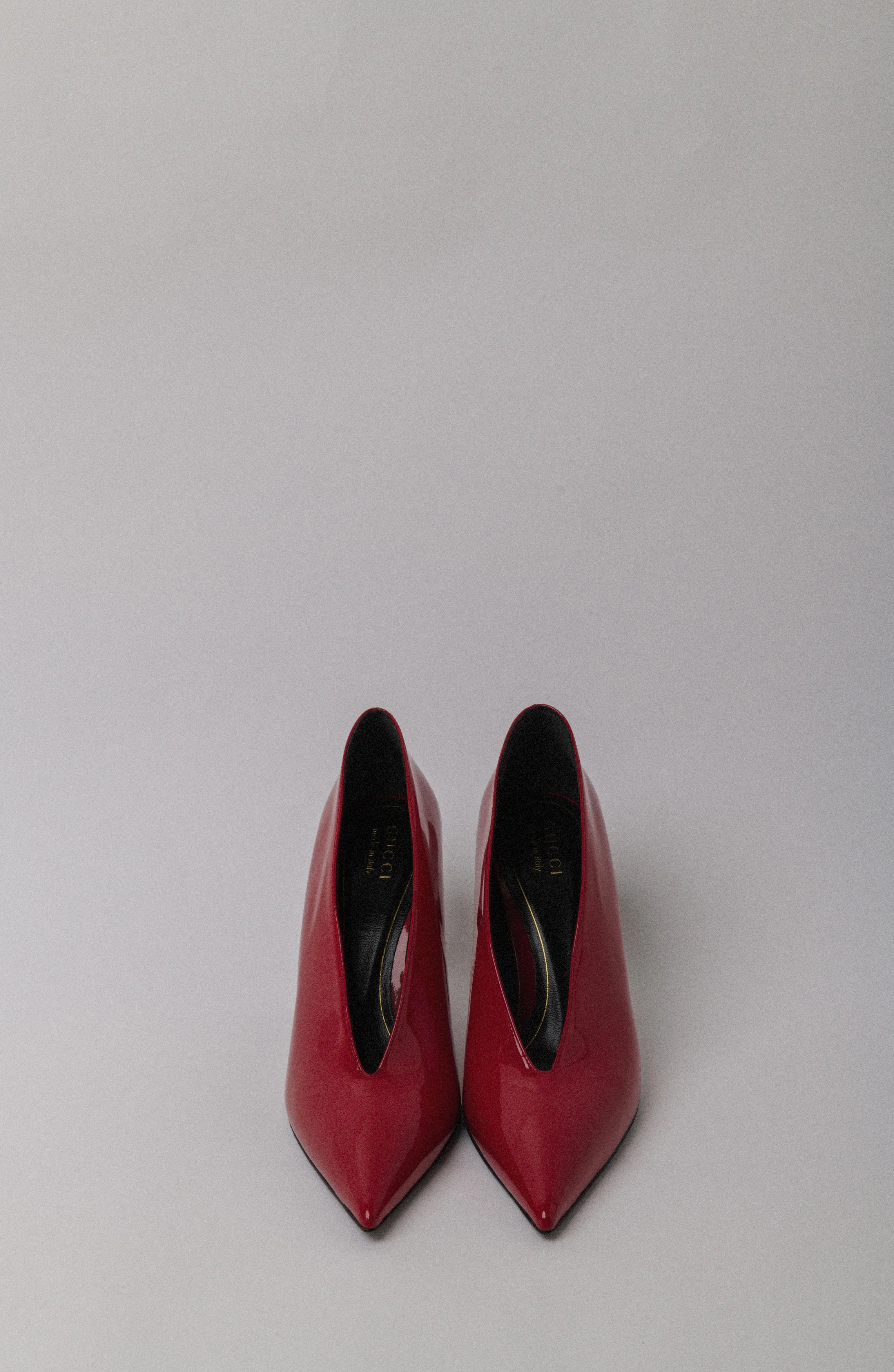 A pair of shiny red high-heeled shoes with pointed toes on a light gray surface.