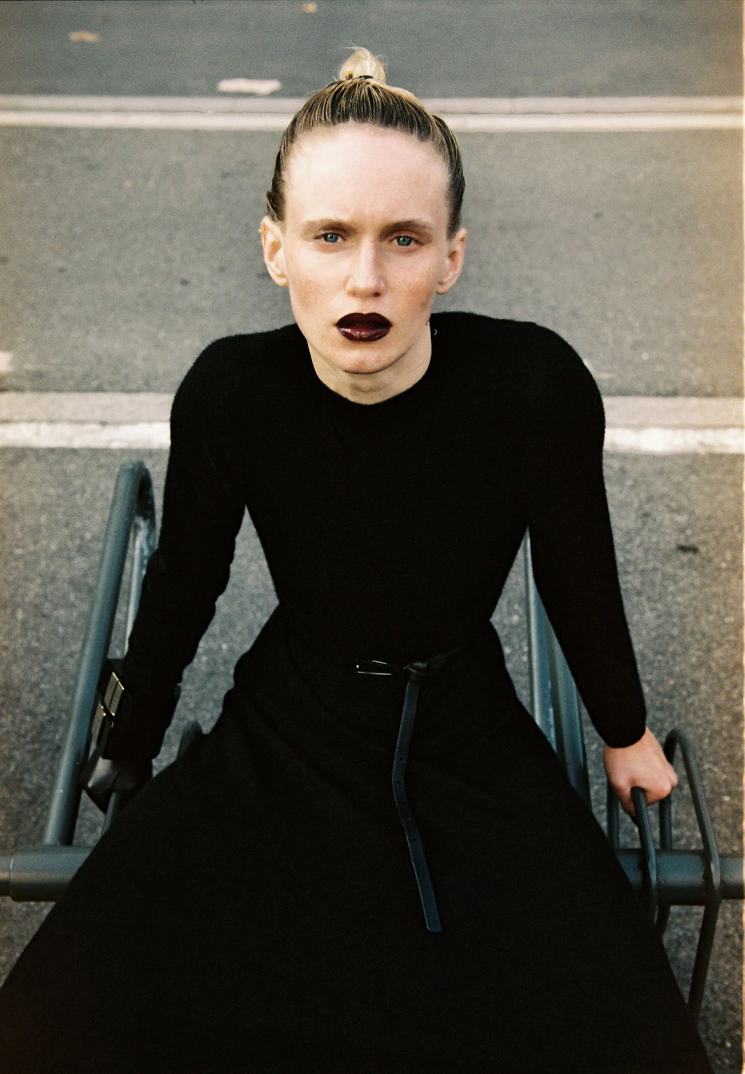 A woman sitting in a wheelchair on a street with painted crosswalk lines, wearing a black long-sleeve top and dark lipstick, looking directly at the camera.
