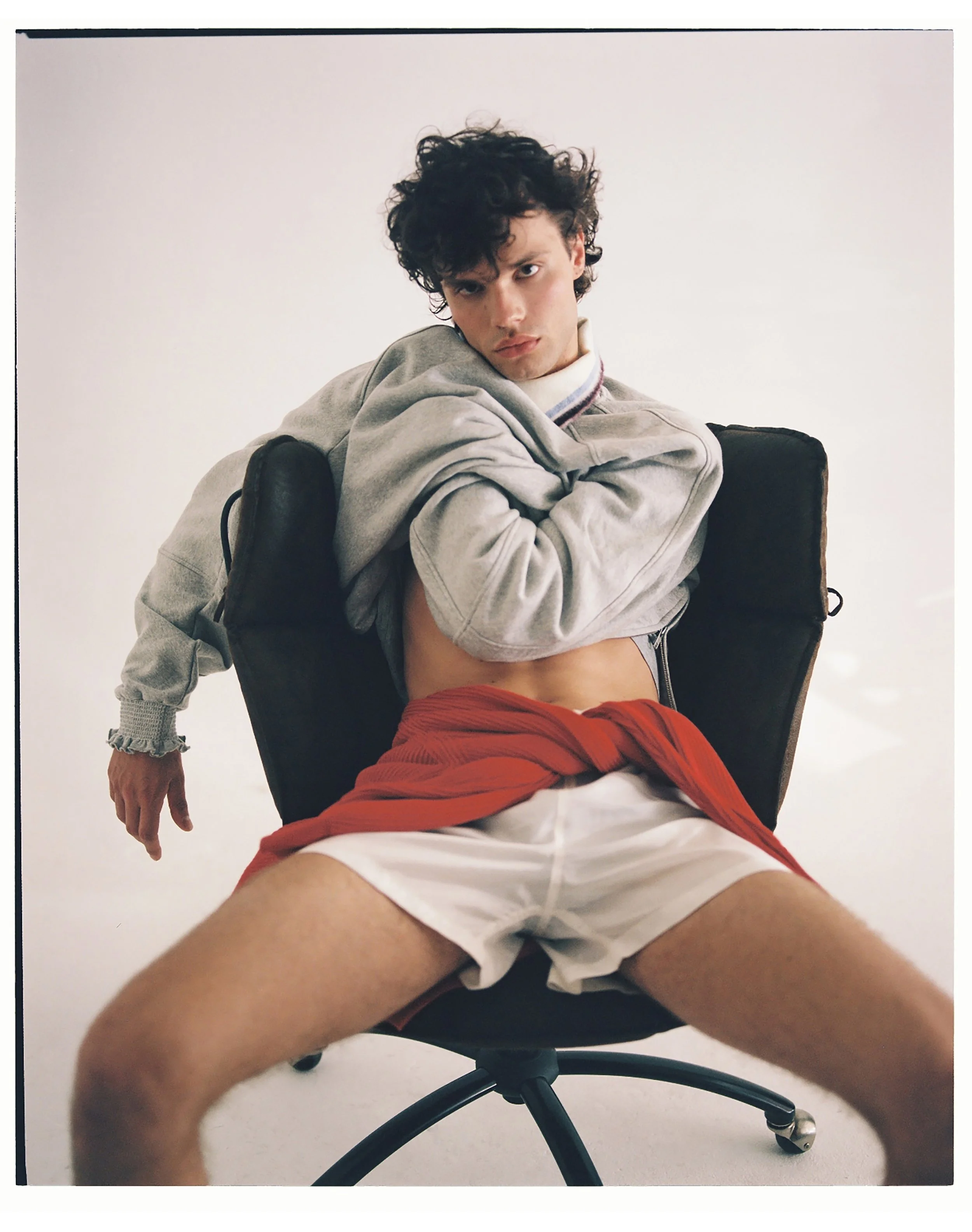 A young man with curly hair sits in an office chair with his legs spread wide apart. He is wearing a gray hoodie, white shorts, and has a red jacket tied around his waist. He is leaning back slightly and has a serious expression on his face.