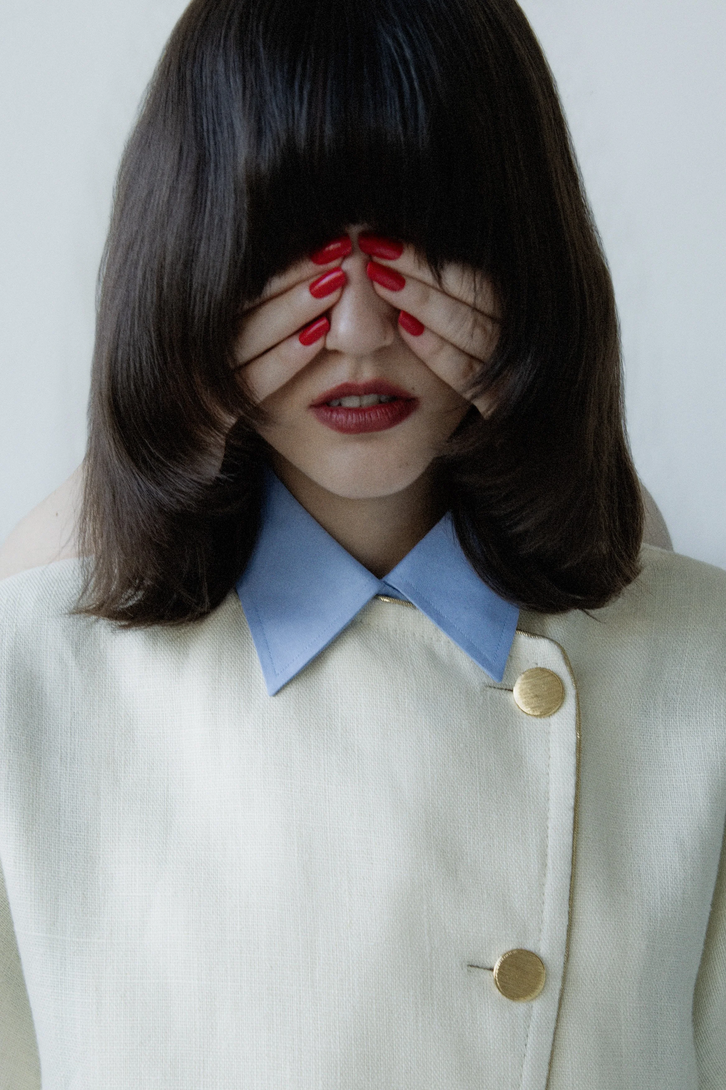 Woman with dark hair and red lipstick covering her eyes with her hands, which have red nails, wearing a cream-colored coat with blue collar and gold buttons.