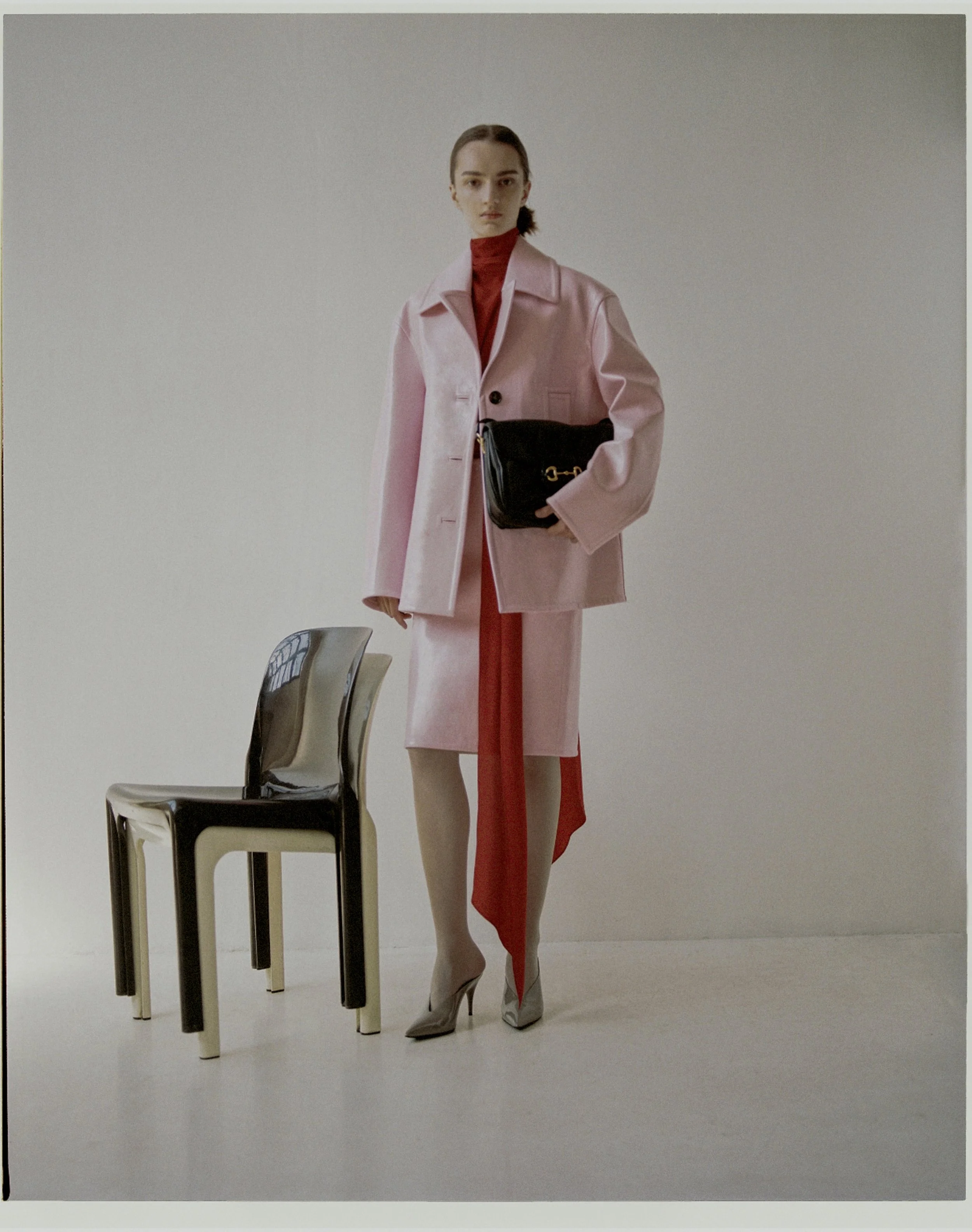 A woman in a pink coat and matching skirt, with a red top underneath, stands next to two chairs, holding a black handbag. She is wearing gray high heels and is positioned against a plain light-colored wall.