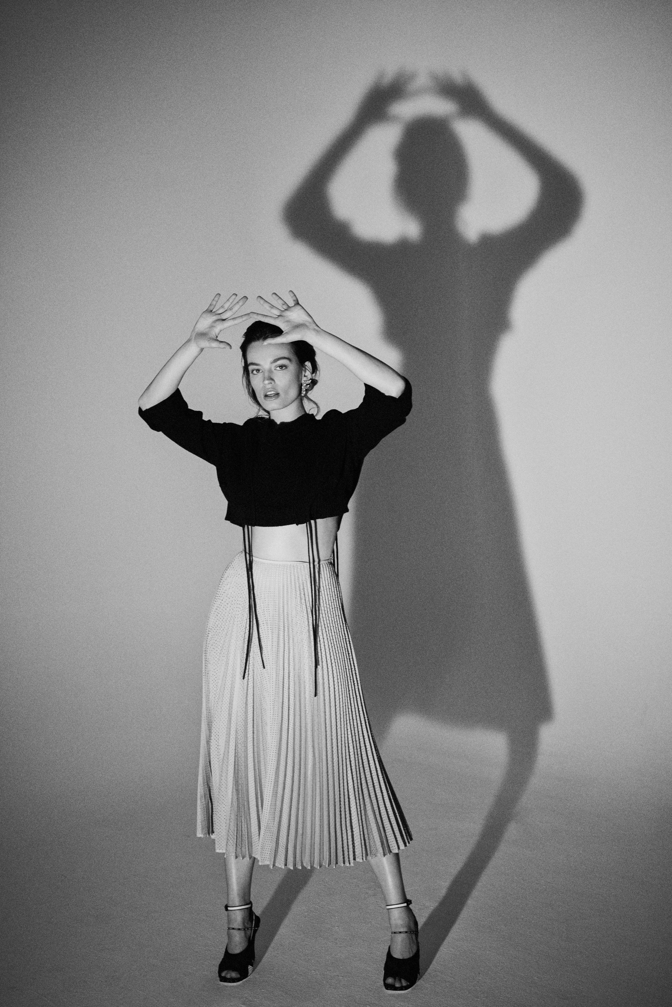 A woman poses with her hands above her head, standing in front of a wall, casting a shadow that resembles a larger figure with arms raised.
