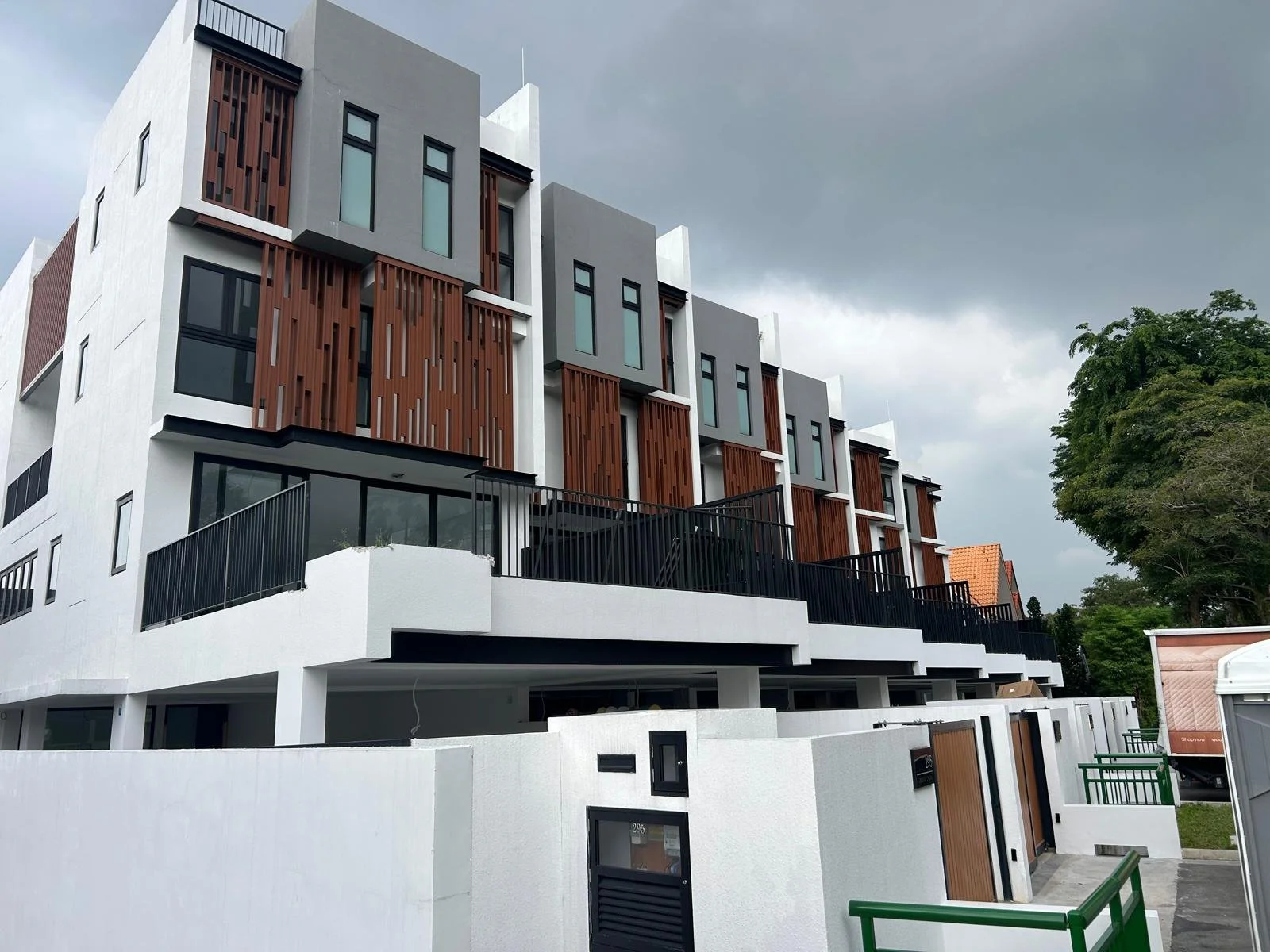 Modern multi-story residential building with white exterior walls, wooden accents, and large glass windows. The building has black railings and a multi-level terrace area. There are trees and a cloudy sky in the background.