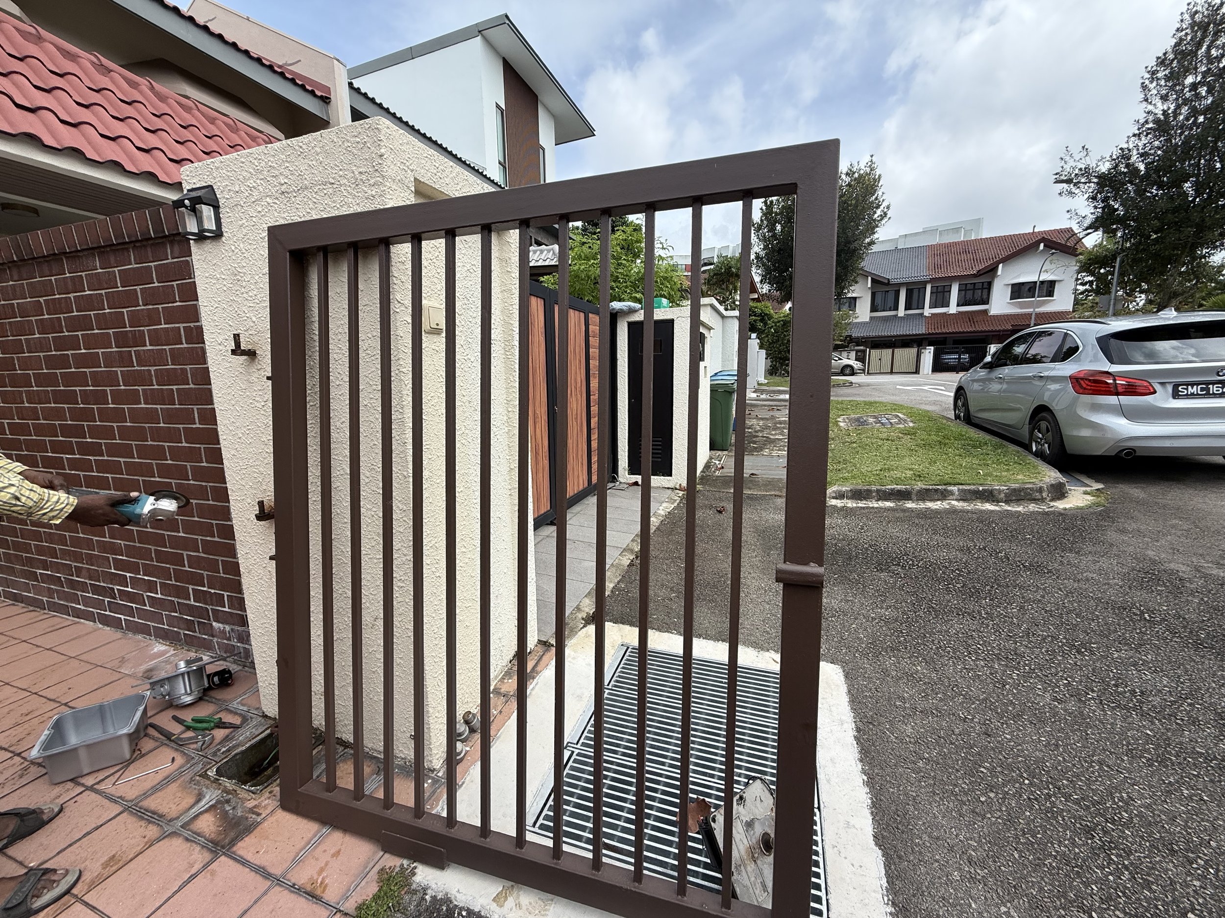 A residential house exterior with a swinging brown metal gate open, revealing a small outdoor area and a walkway leading to the front door, with parked cars and other houses visible in the background.