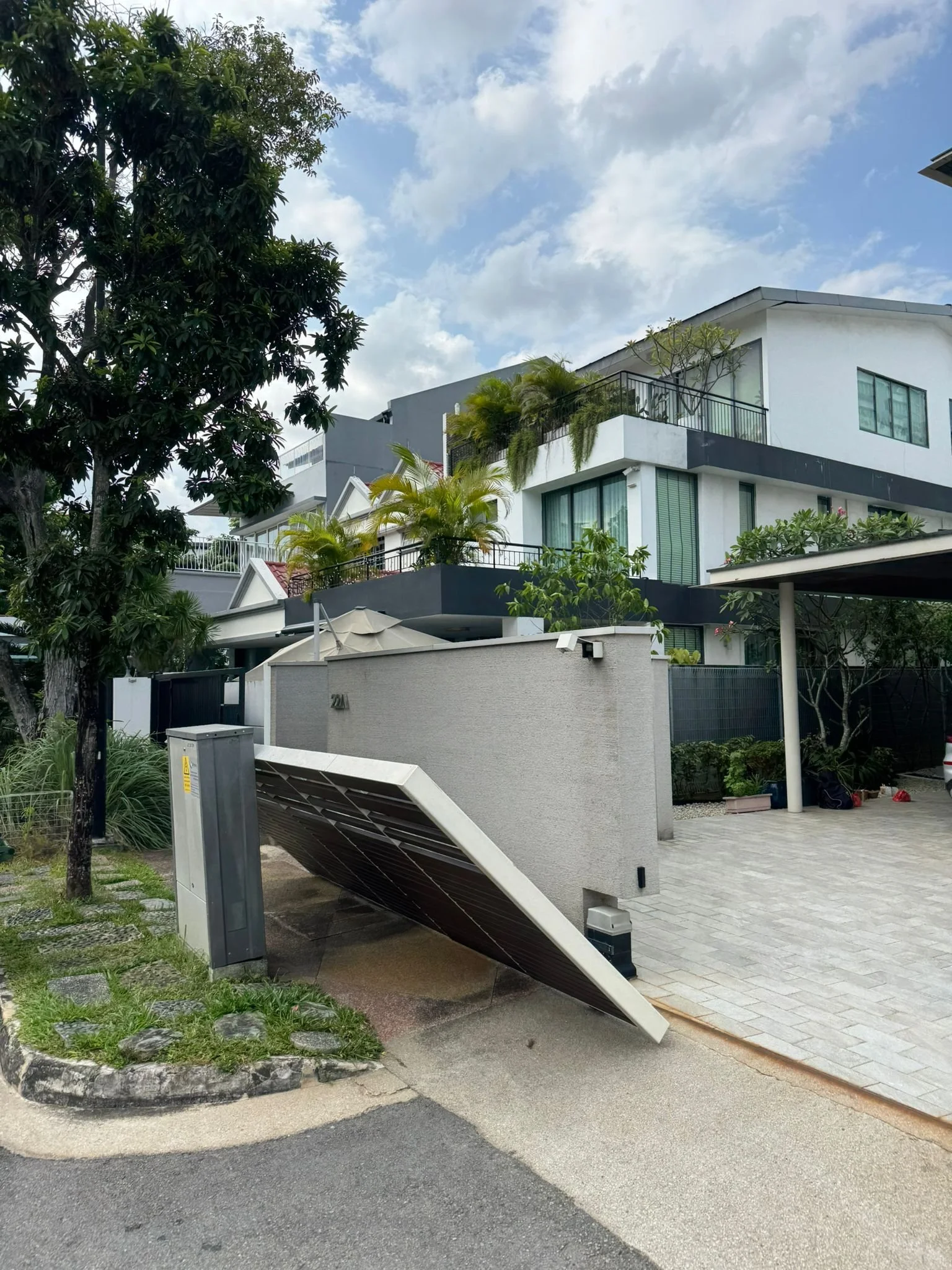 A modern multi-story house with balconies, lush green plants, and a solar panel on the driveway. The house has white and black exterior walls and glass doors. A tree and cloudy sky are visible in the background.