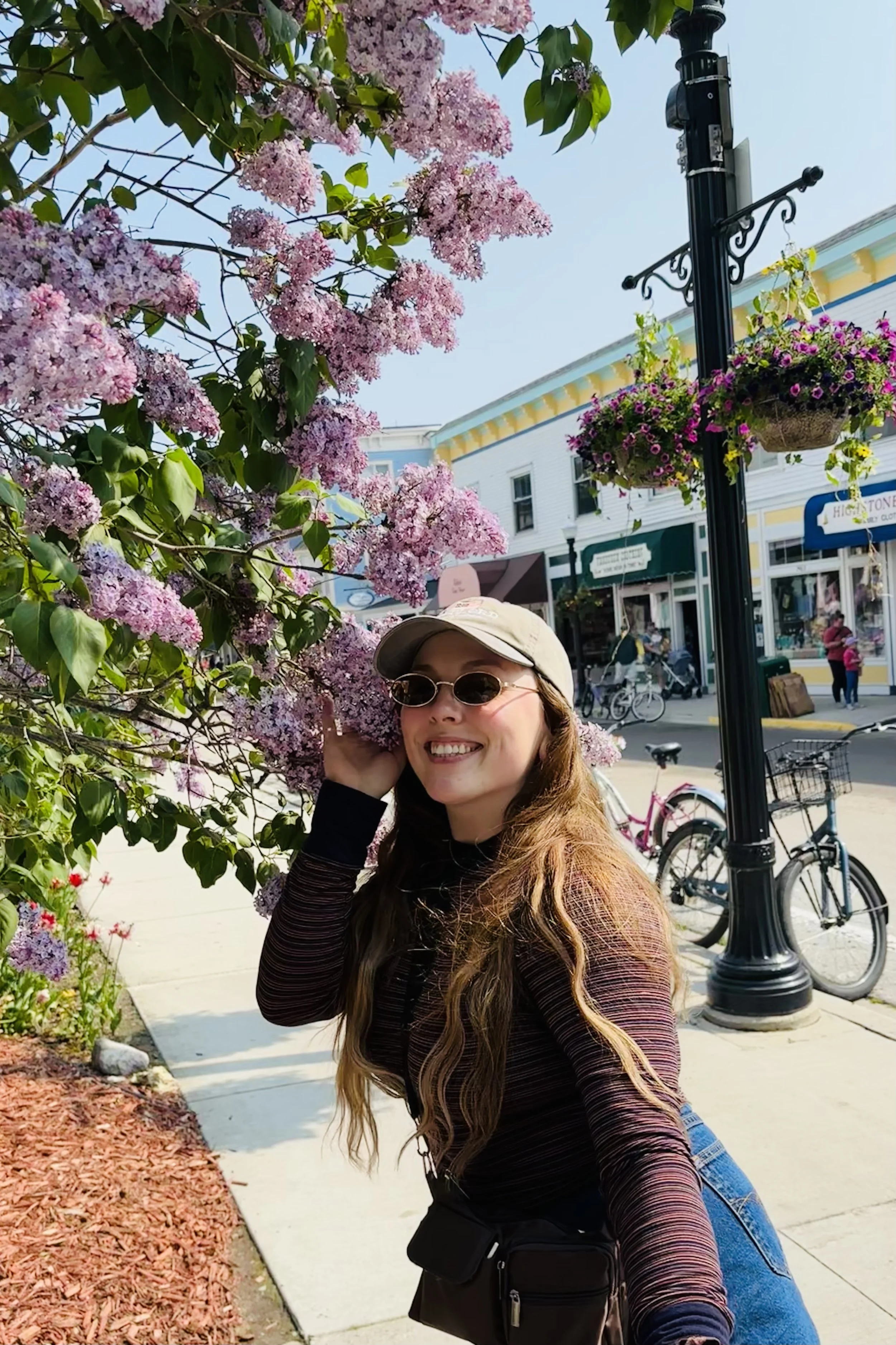 A woman with long wavy hair wearing sunglasses, a beige cap, and a black jacket smiles while standing near pink flowering bush on a sidewalk with bicycles and storefronts in the background.