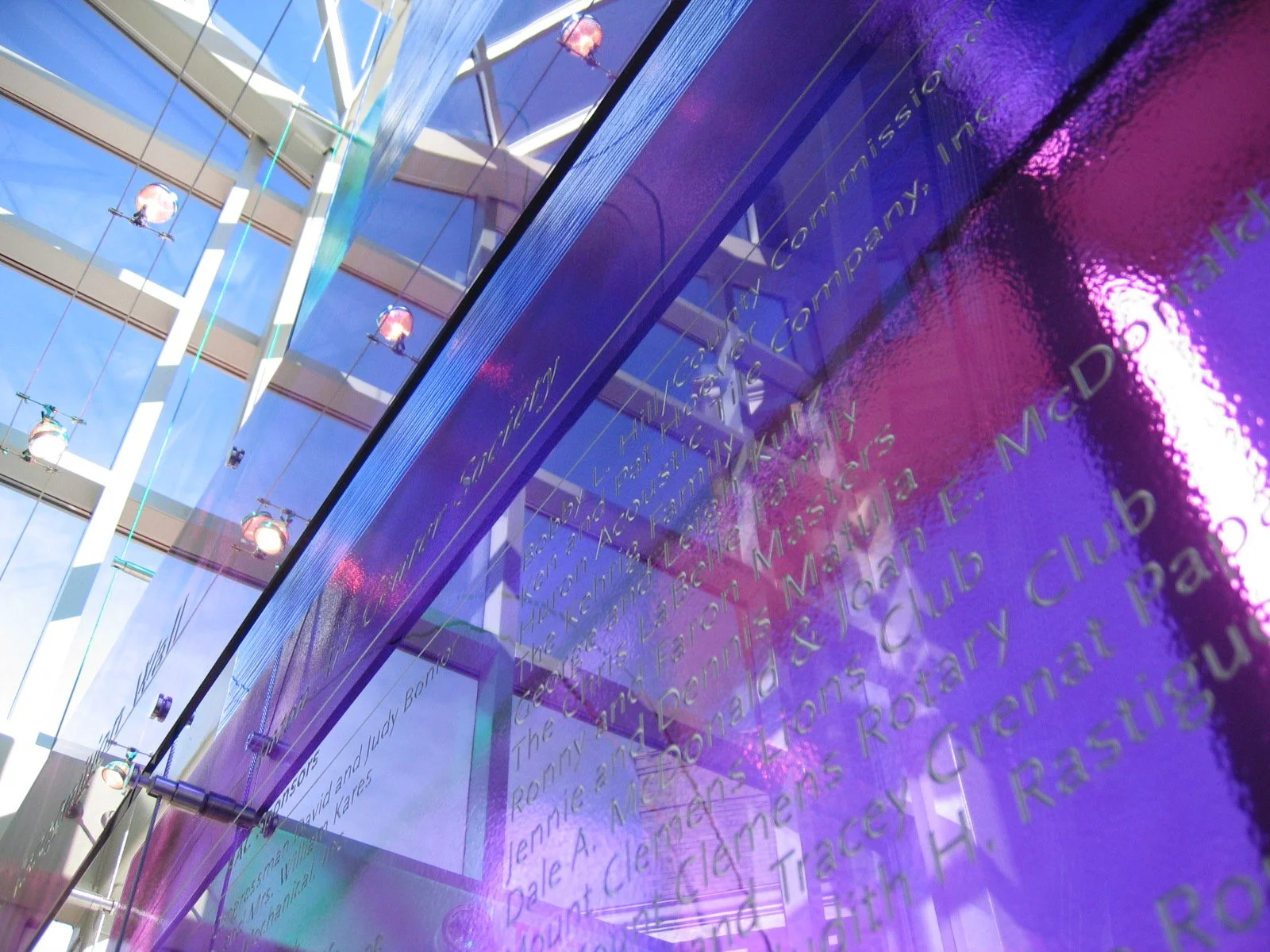 Interior of a modern building with a glass roof and walls, reflecting a purple illuminated wall with gold script writing. The reflection shows a striped white and brown roof structure with hanging lights.