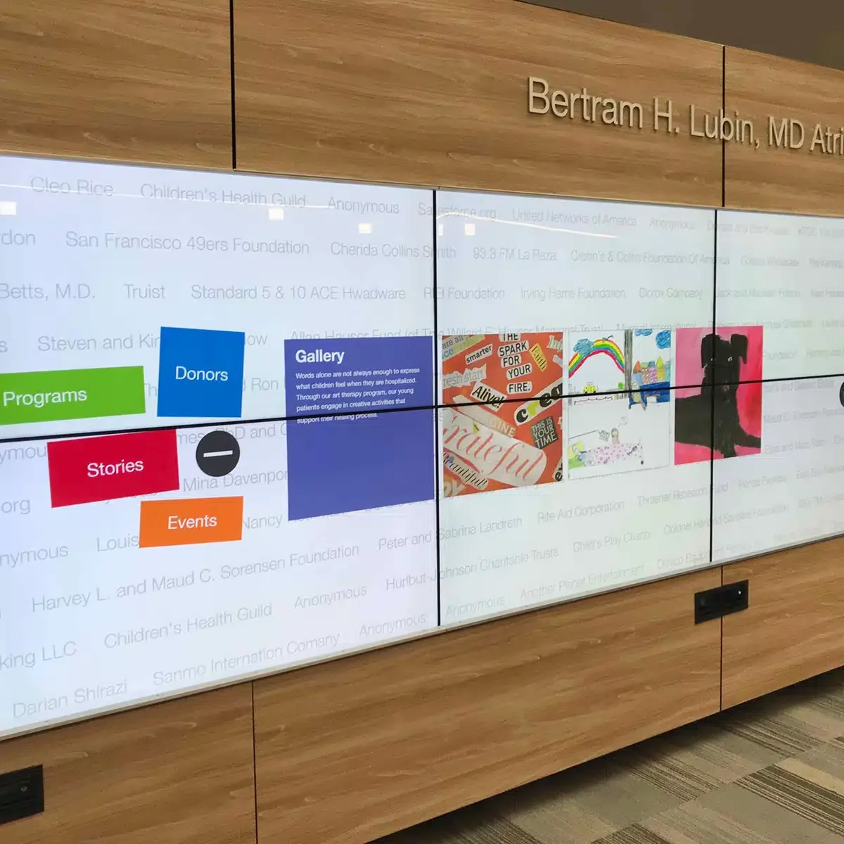 Multisection digital display board with colorful icons and text, mounted on a wood-paneled wall, showing various programs and donor information.