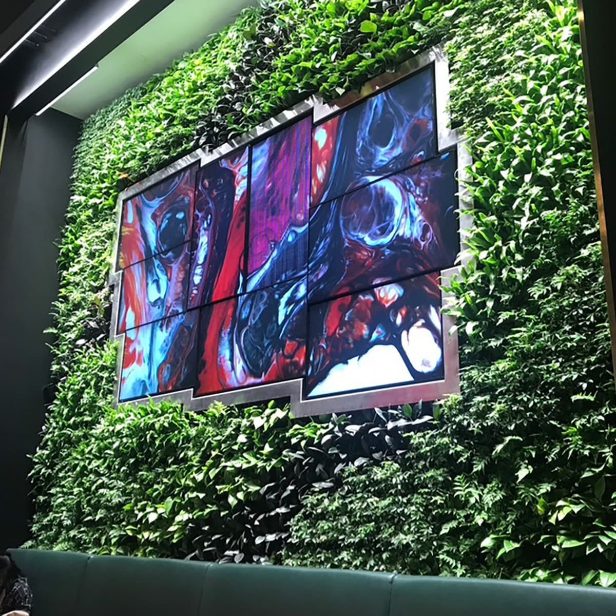 A digital artwork with abstract swirling colors in red, black, white, and blue, mounted on a wall covered with green foliage, with a dark sofa in the foreground.