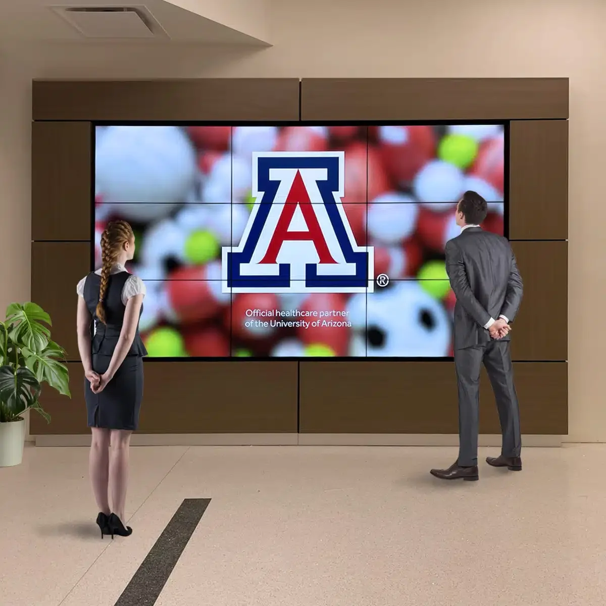 Two people, a woman and a man, standing in front of a large screen displaying the University of Arizona logo with a background of colored golf balls. The woman has long braided hair and is dressed in a professional outfit, while the man is in a suit. There is a potted plant on the left side.