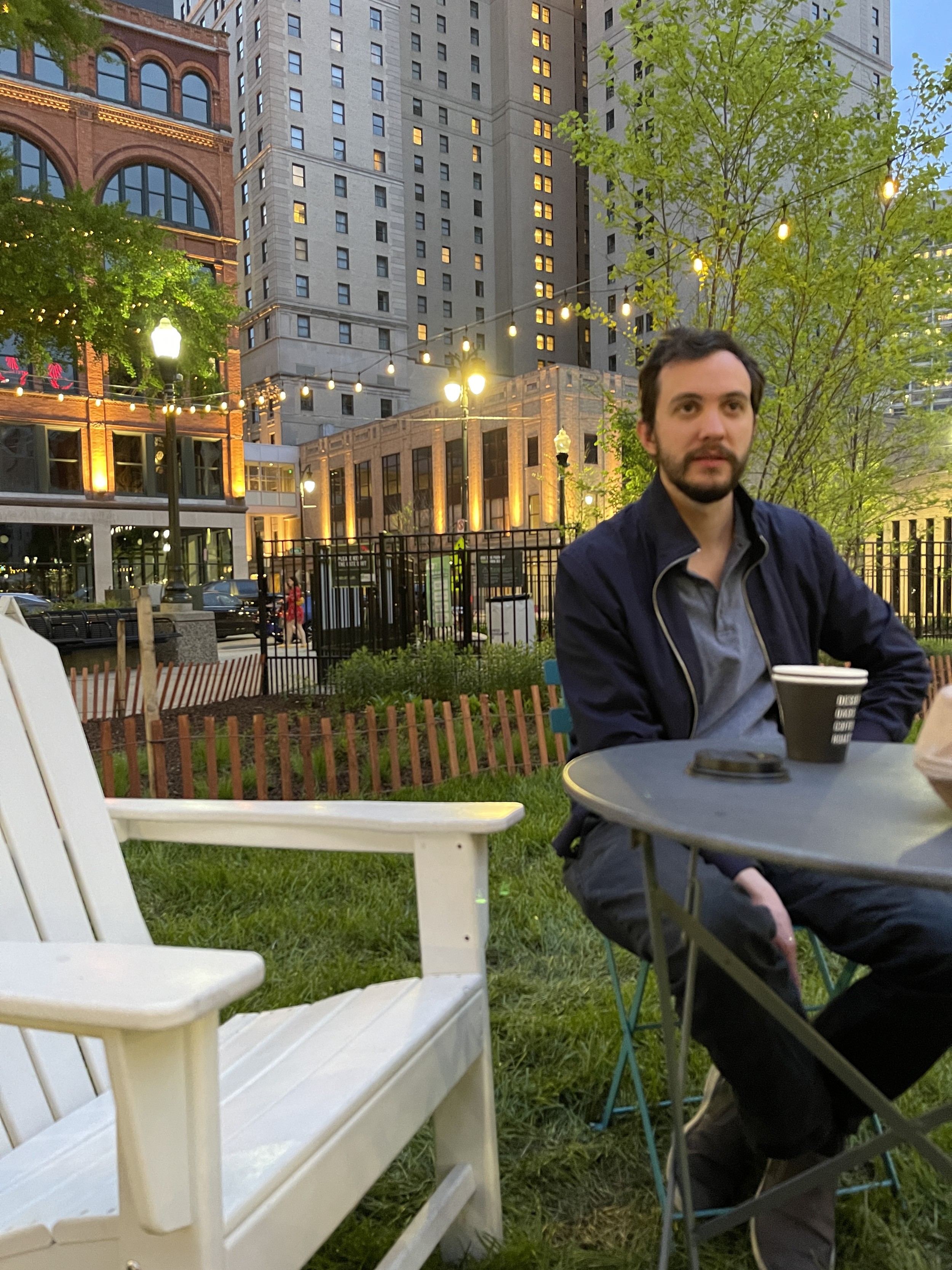 A man sitting at an outdoor table in an urban park at dusk, with city buildings, string lights, and trees in the background.