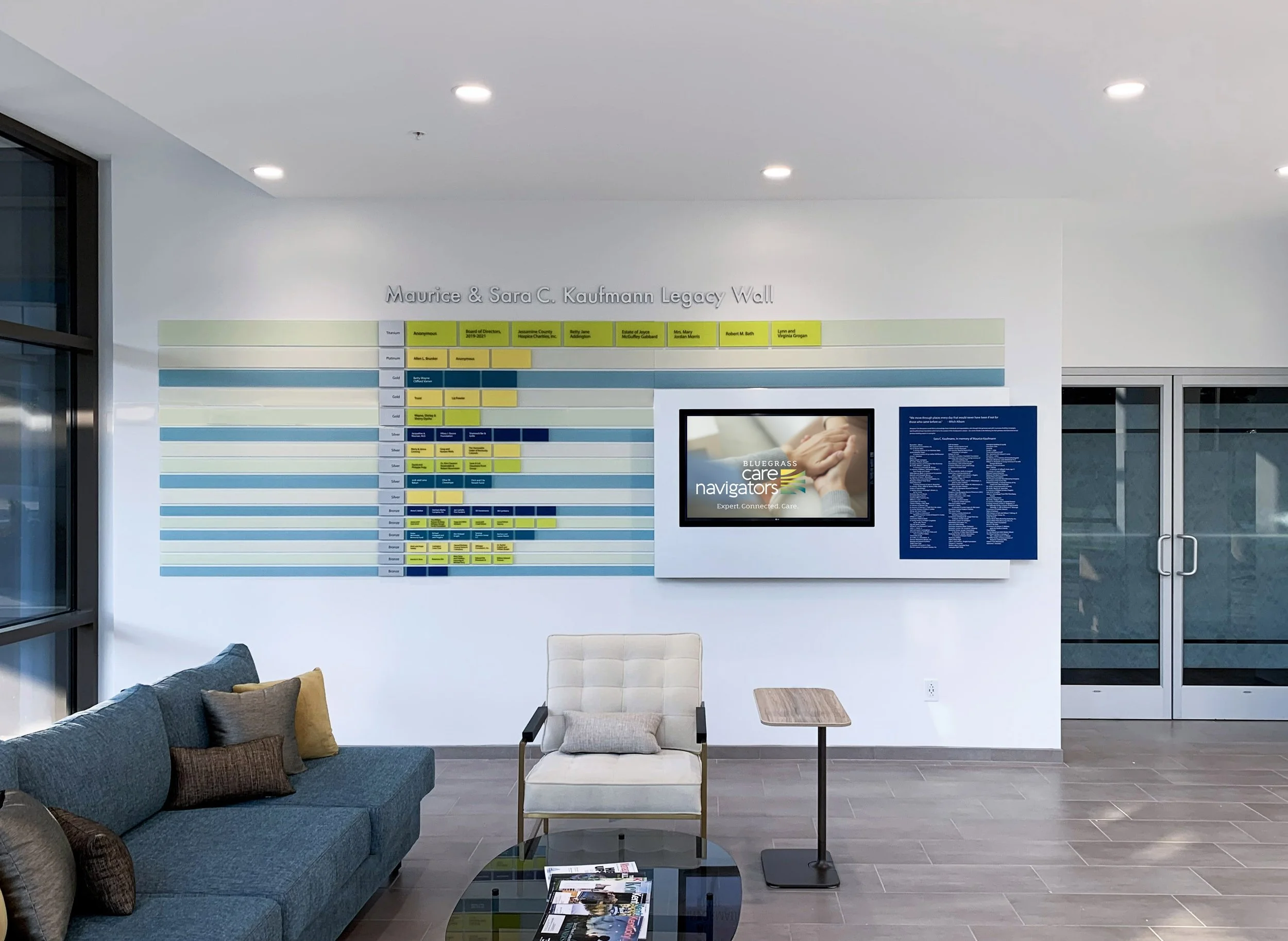 Lobby area with a seating sofa and chair, a small side table, a wall-mounted digital screen displaying 'Bluegrass Care Navigators,' and a Wall of Honor titled 'Maurice & Sara C. Kaufmann Legacy Wall' with various colored name plaques.