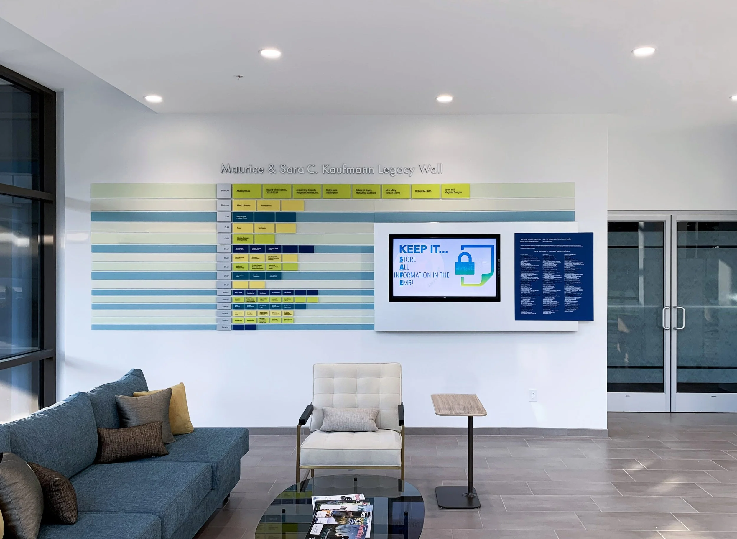 Lobby area with a wall display titled 'Maurice & Sara C. Kaufmann Legacy Wall,' a digital screen with a message about storing information, a seating area with a blue sofa with pillows, a white chair, a small table, and glass double doors.