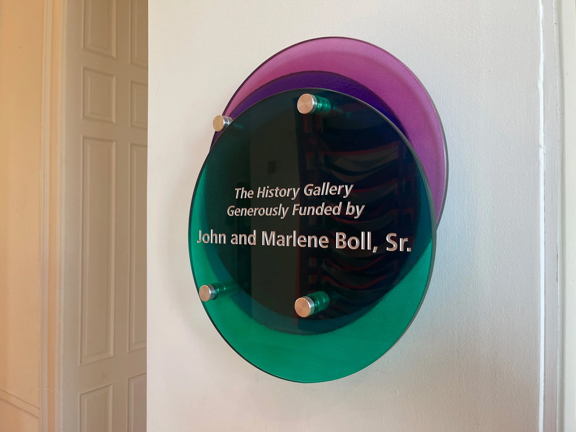 A circular decorative plaque mounted on a wall with text that reads 'The History Gallery Generously Funded by John and Marlene Boll, Sr.' The plaque has a layered, colorful design with purple, green, and dark shades.