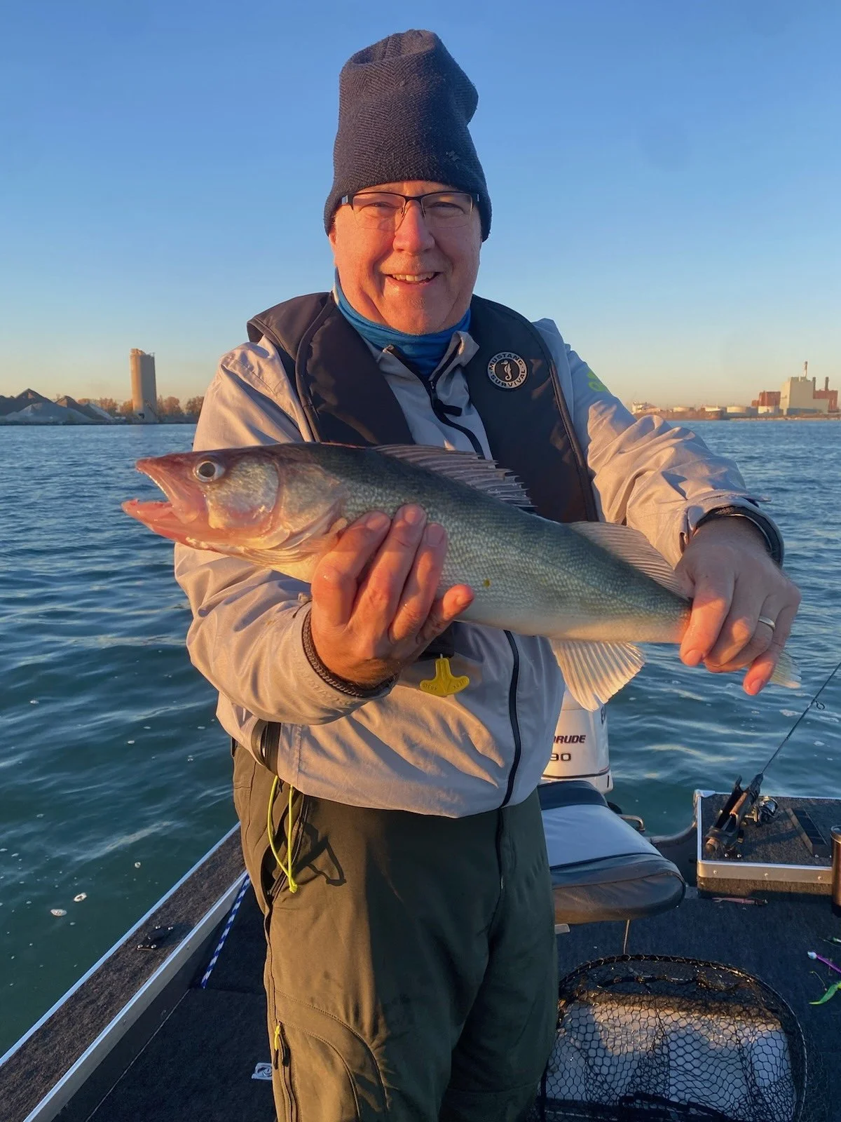 Man wearing a black beanie and glasses holding a large fish on a boat with water and cityscape in the background.