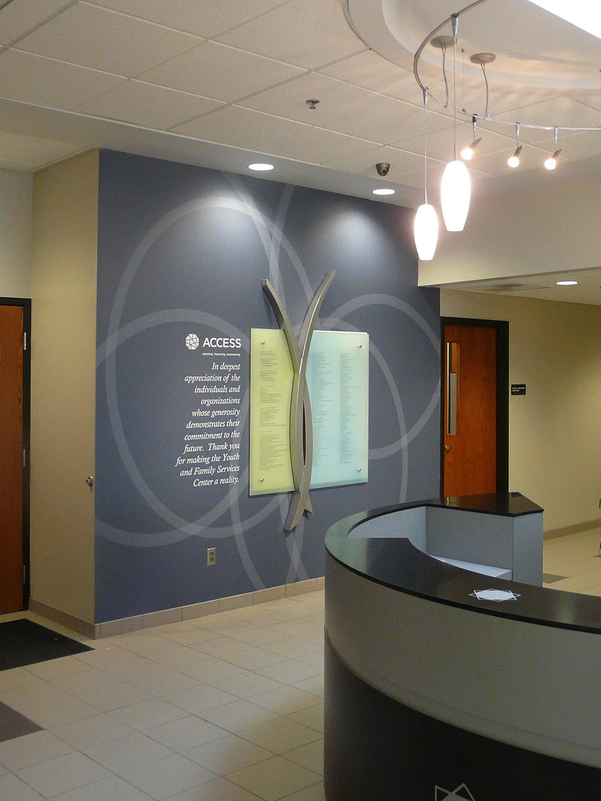 Lobby with a blue accent wall featuring a decorative metal art piece and a sign expressing appreciation for donors, in front of a reception desk with a curved counter, illuminated by hanging lights.