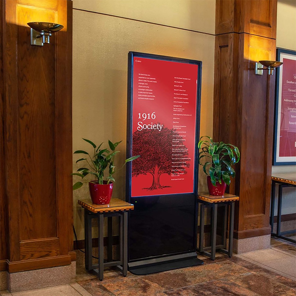 A digital display sign with the text '1916 Society' and a red background, flanked by two potted plants on small tables, in a wood-paneled interior.