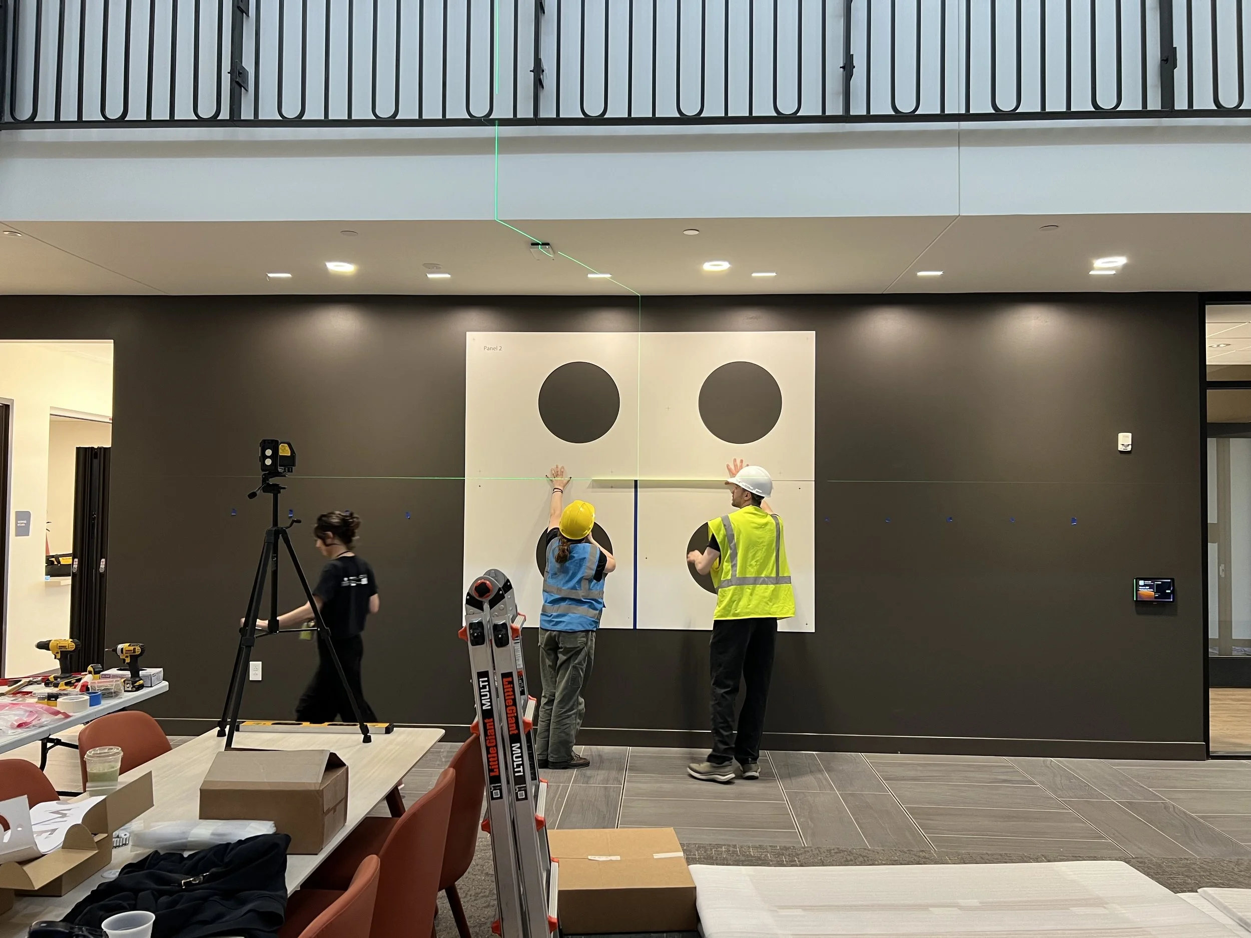 Workers installing or inspecting a large wall graphic with two black circles on a white background in an indoor space, with tools and equipment on nearby tables.