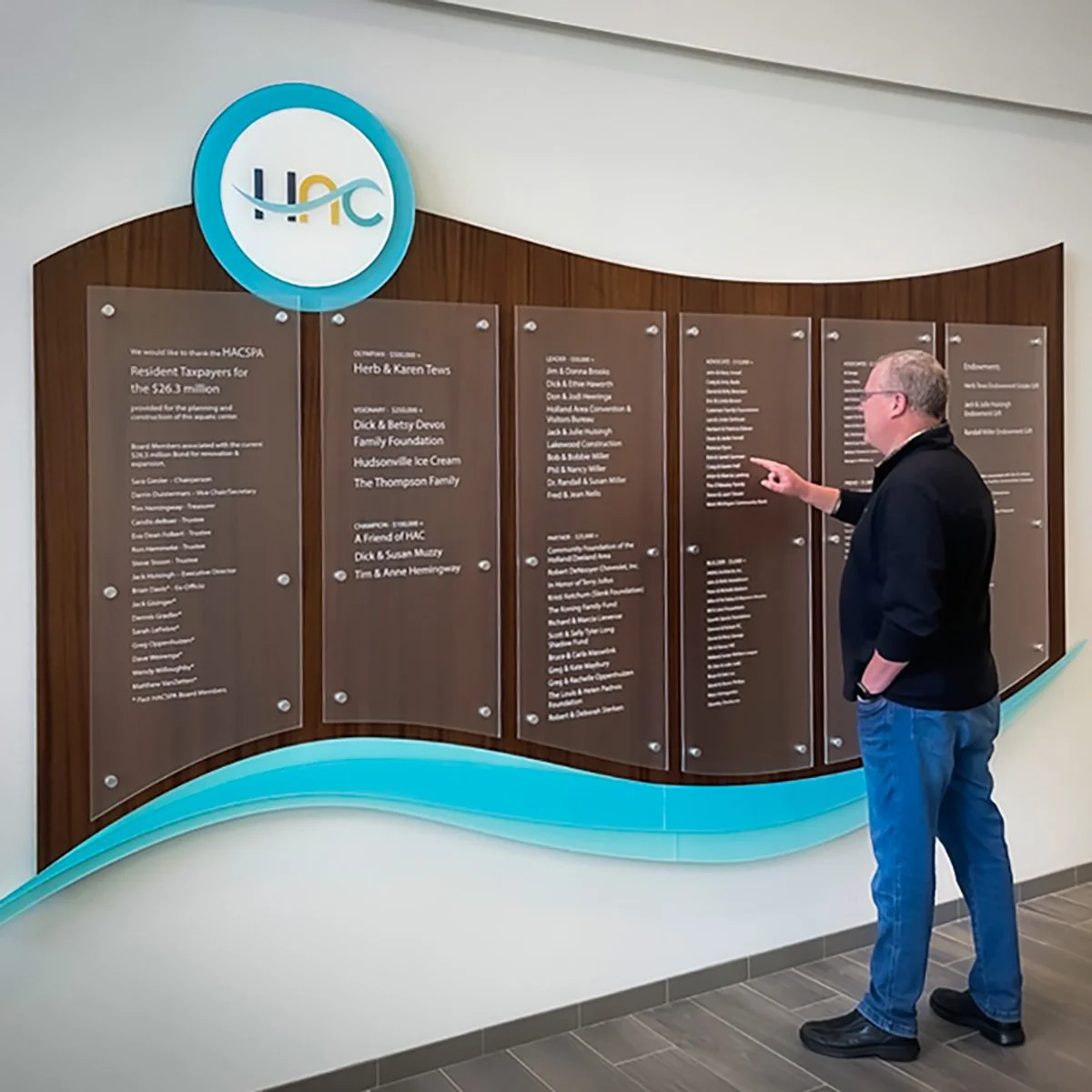 A man in glasses, a black jacket, and blue jeans points at a large wall display with multiple inscribed panels and an HACC logo at the top.