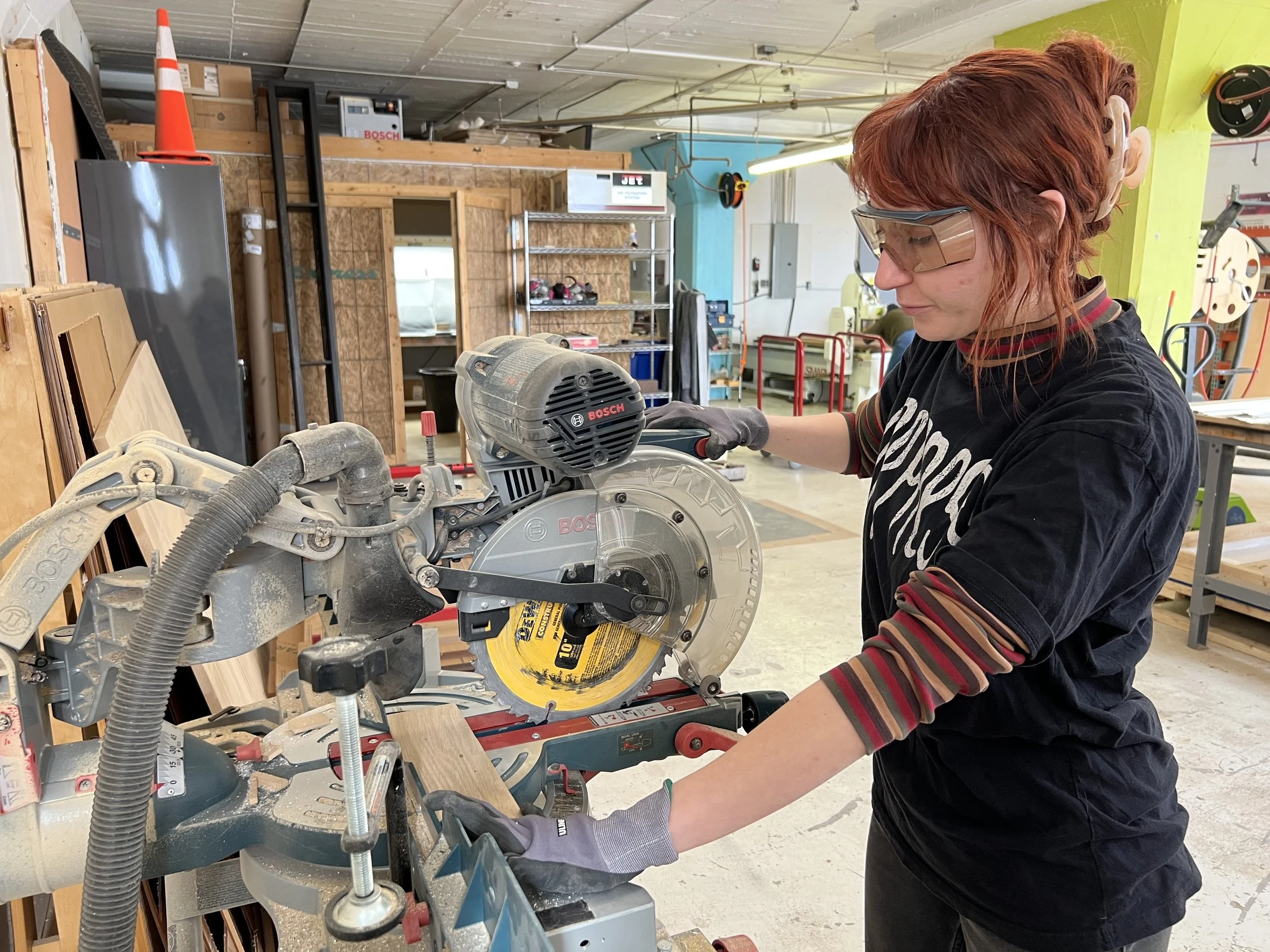 A woman in safety glasses and gloves using a Bosch miter saw to cut wood in a woodworking shop.