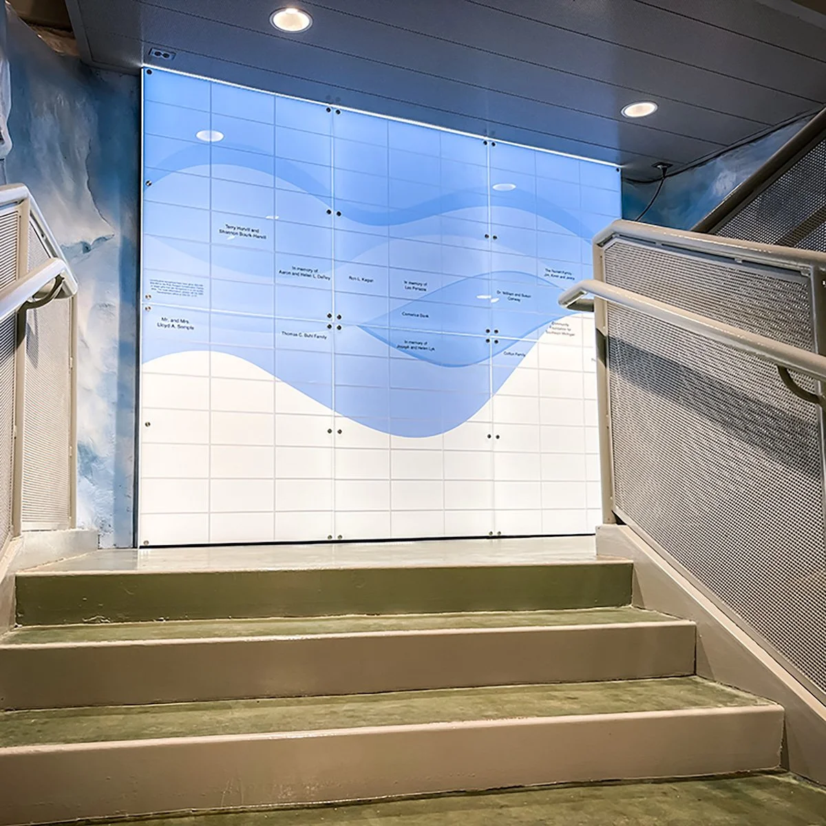 A staircase leading up to a wall-mounted memorial plaque with a blue wave design, dedicated to Tony Marshall and Shannon Dela Rosa, with mentions of Alexit A. Springer, Thomas C. Bush Family, and others, in a public indoor space.