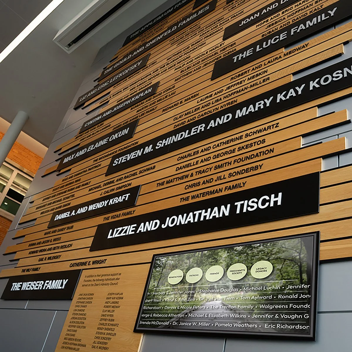 A large wall display with black and wood-colored panels listing names of families and donors at a public building, possibly a hospital or community center.