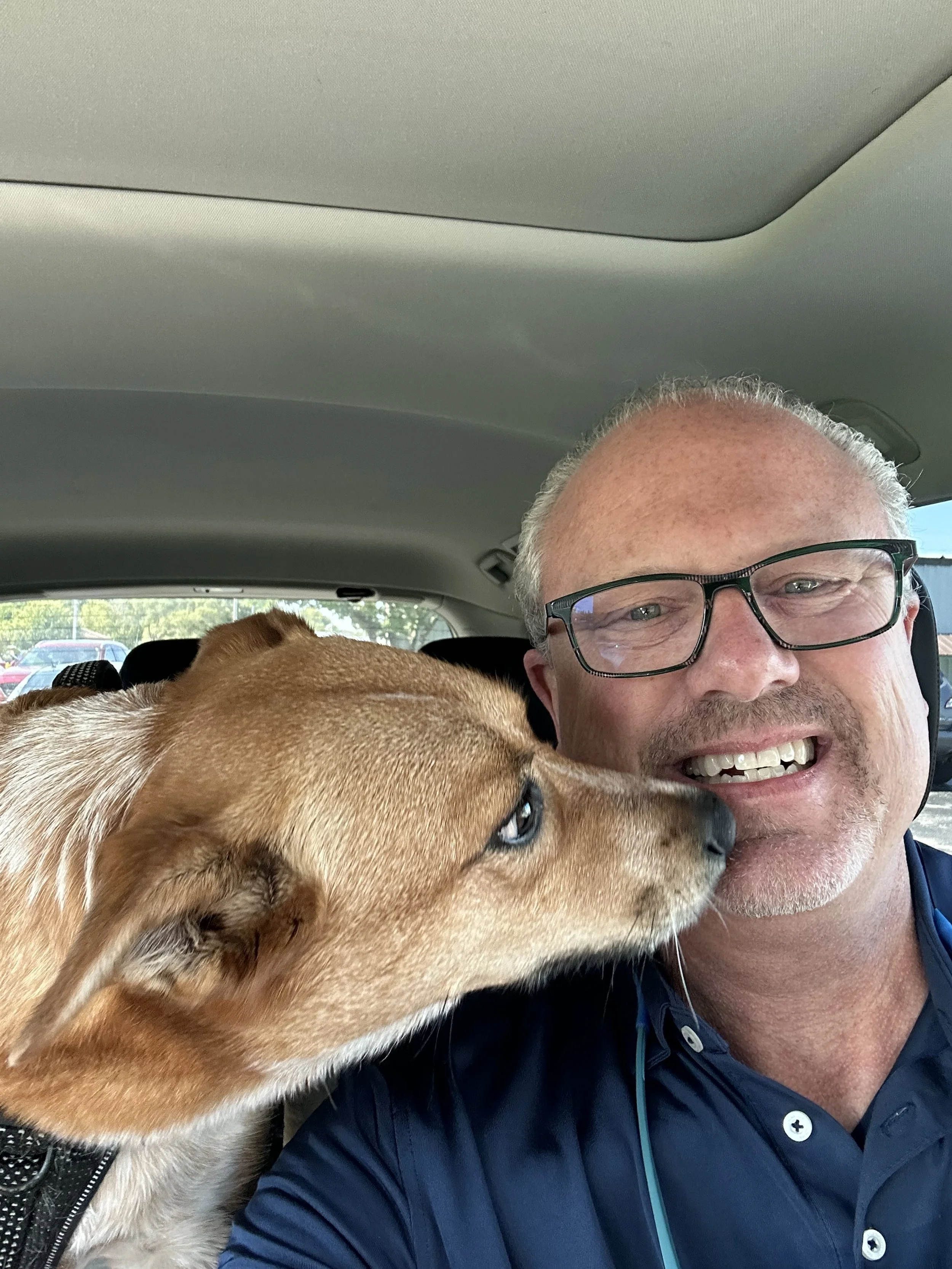 A man smiling with a dog nuzzling his face inside a car.