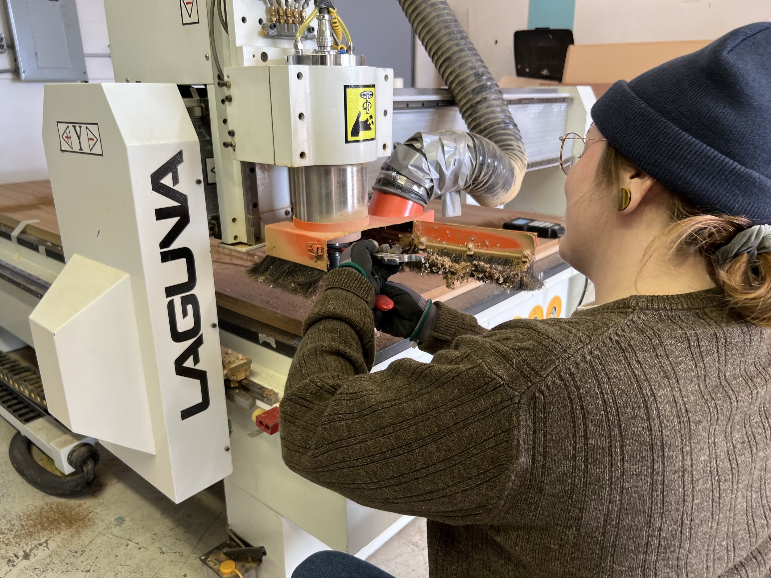 A woman wearing glasses, a dark beanie, and a brown sweater operating a saw machine in a workshop. The machine is branded 'LAGUNA' and is cutting a piece of material, producing sawdust and debris.