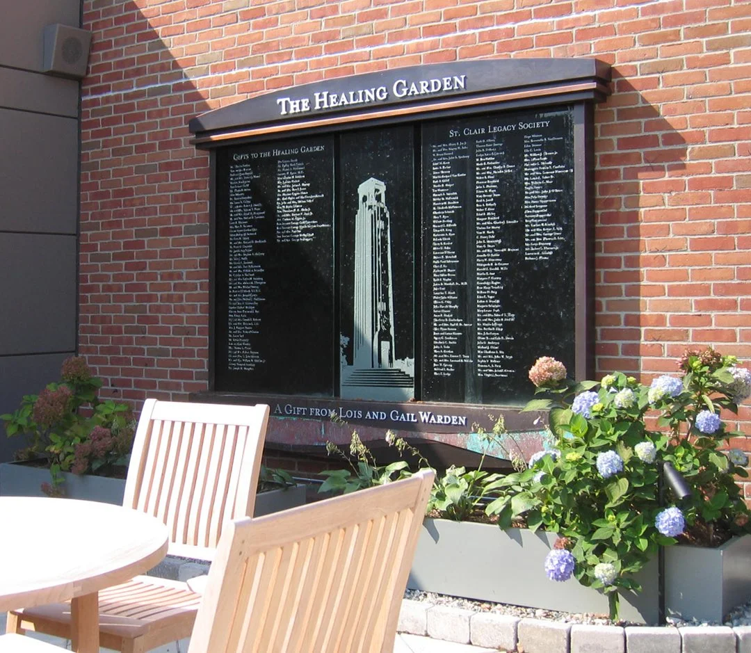 A memorial plaque titled 'The Healing Garden' mounted on a brick wall, with engraved names and a depiction of a tower, surrounded by garden beds with flowers and outdoor furniture.
