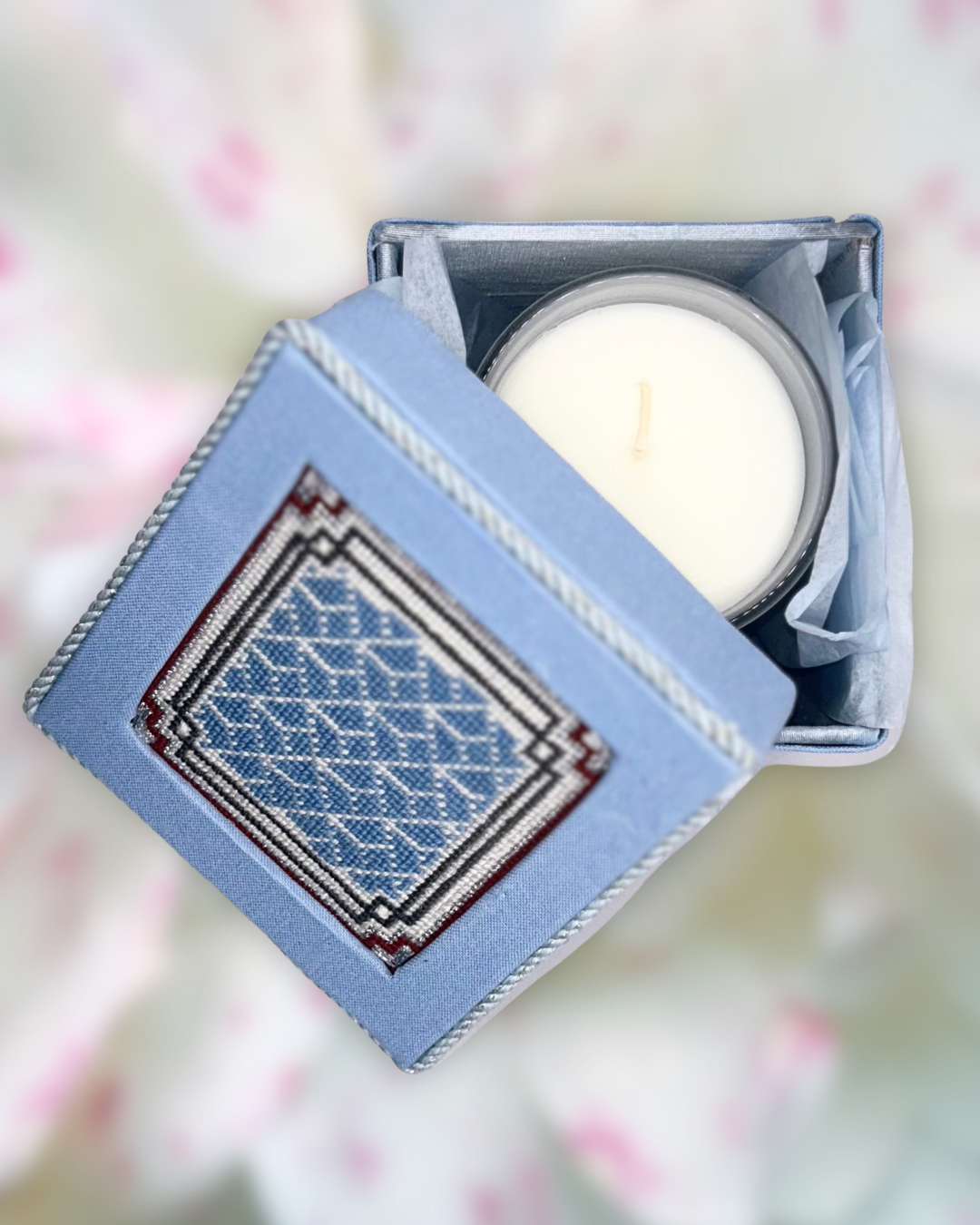 A white candle inside a decorative box with a blue embroidered cover.