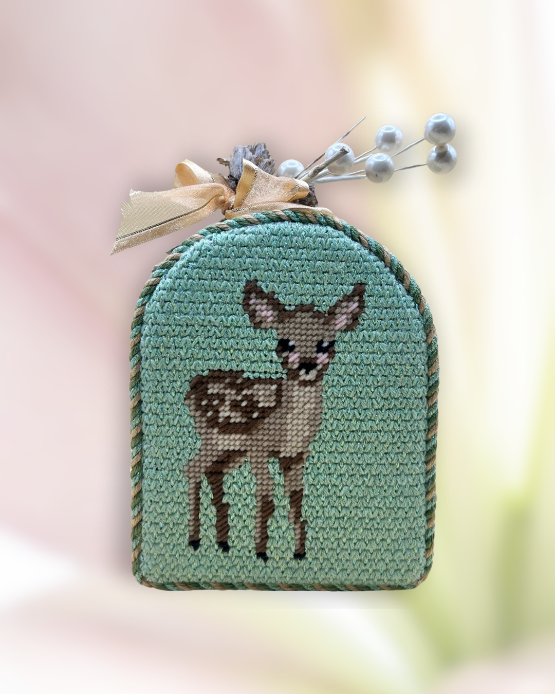 Decorative cross-stitched standup with a deer design, adorned with a beige ribbon and pearled sprig.
